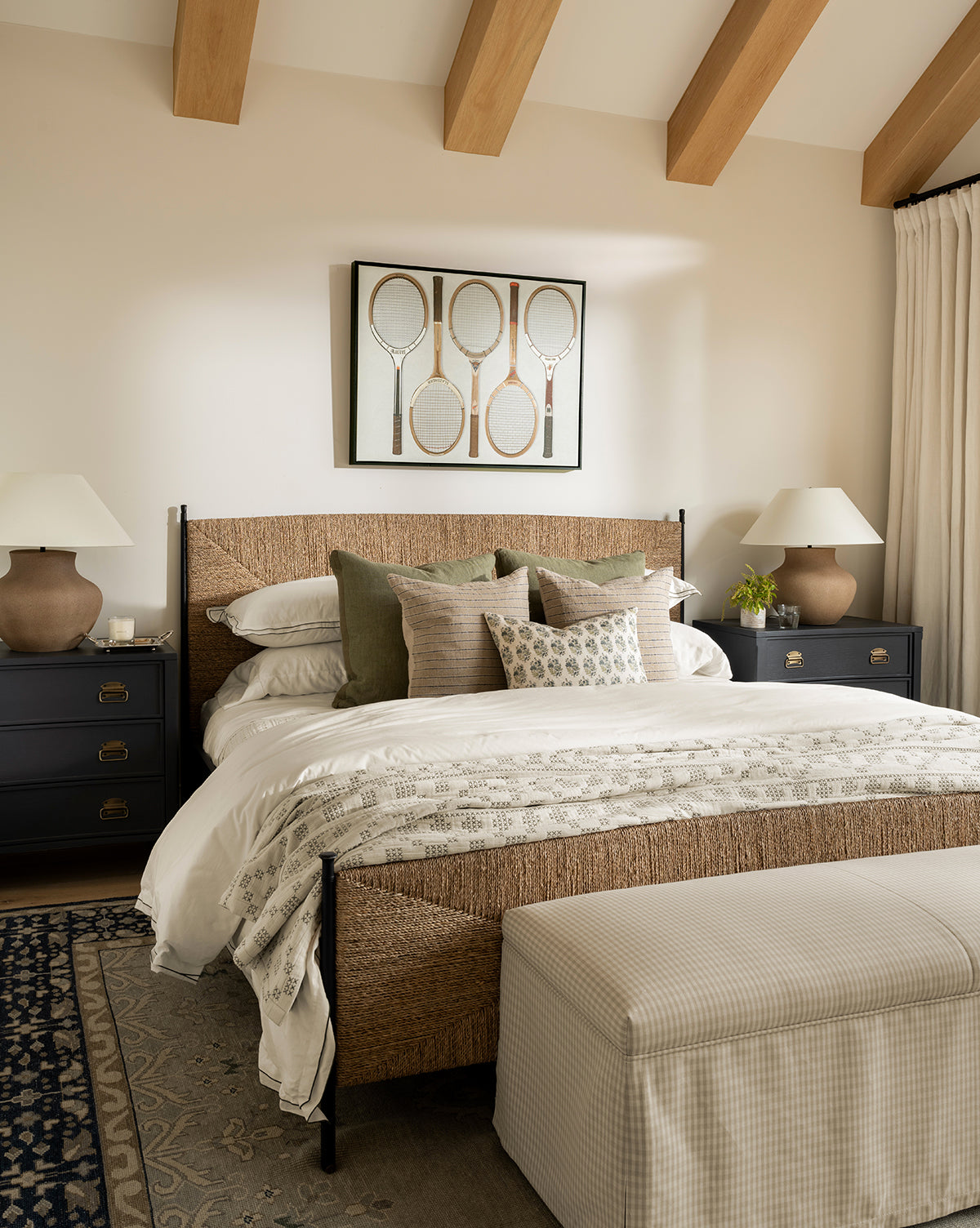 A cozy bedroom featuring the Peregrine Bed with a woven seagrass headboard, layered pillows and blankets, black nightstands with white lamps, exposed wood beams, tennis racket artwork above the bed, and a checkered bench at the foot, by McGee & Co.