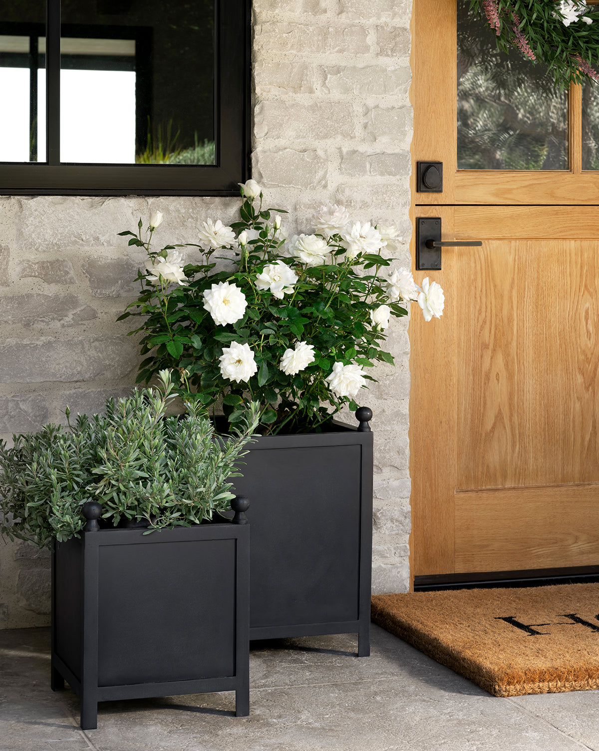 Two black Clariance Planter squares filled with white flowers and greenery sit on a stone porch next to a wooden door with a glass window, beside a tan doormat, all accented by the light gray stone wall for outdoor style.