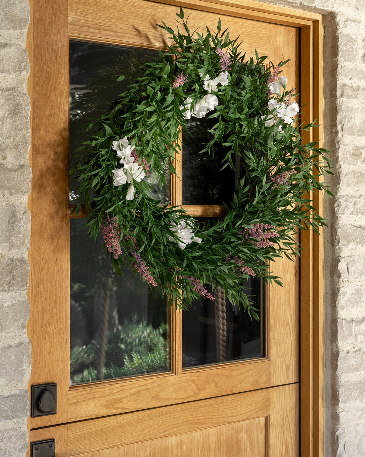 The Faux Bamboo & Sweet Pea Wreath, adorned with green leaves and sweet pea flowers, hangs on a glass-paneled wooden door set in a stone exterior wall, by McGee & Co.
