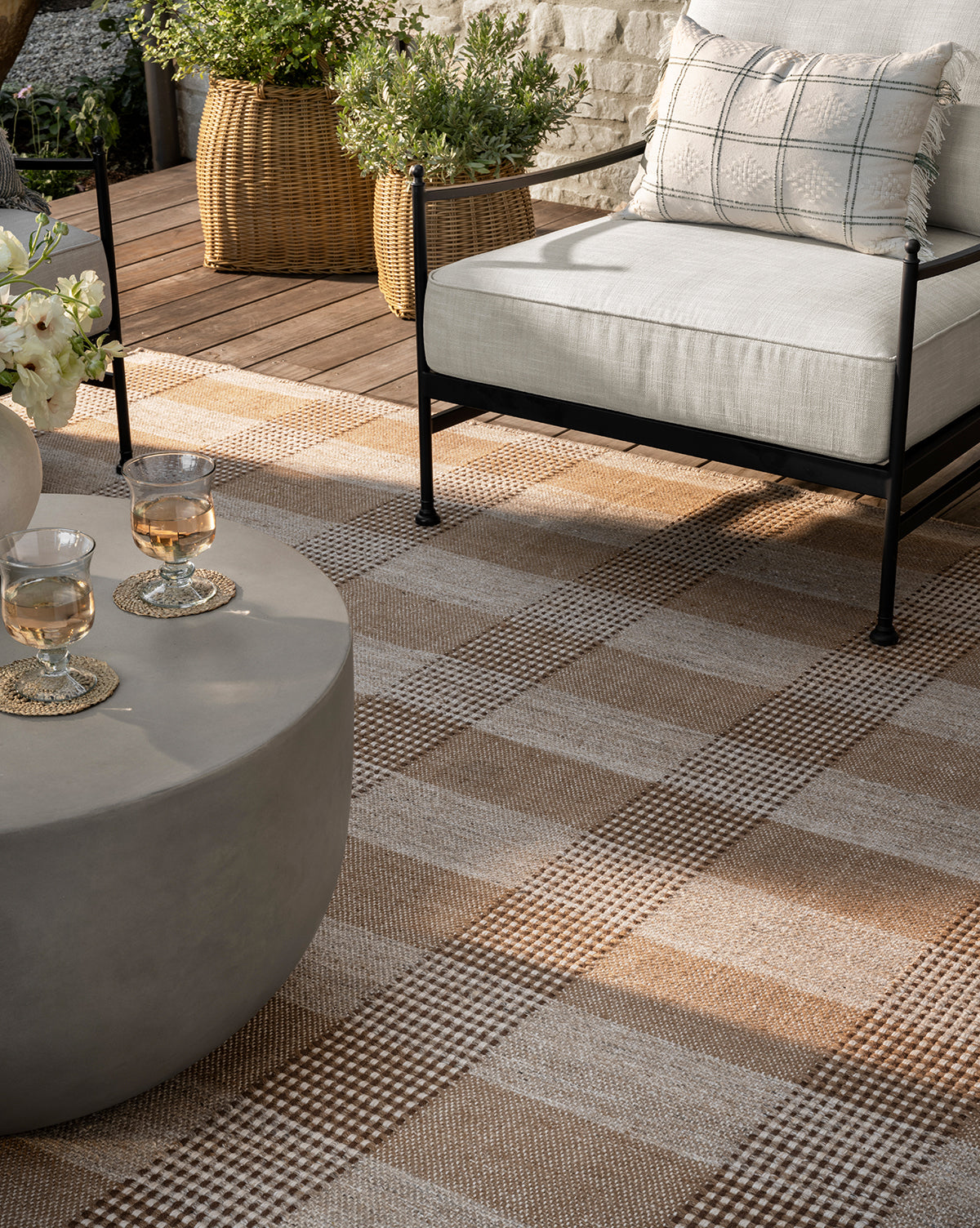 Modern outdoor patio featuring the Hargrove Handwoven Indoor/Outdoor Rug in a beige and brown checker pattern, a cushioned chair with plaid pillow, round gray coffee table with two drinks, and plants in woven baskets on the wooden deck.