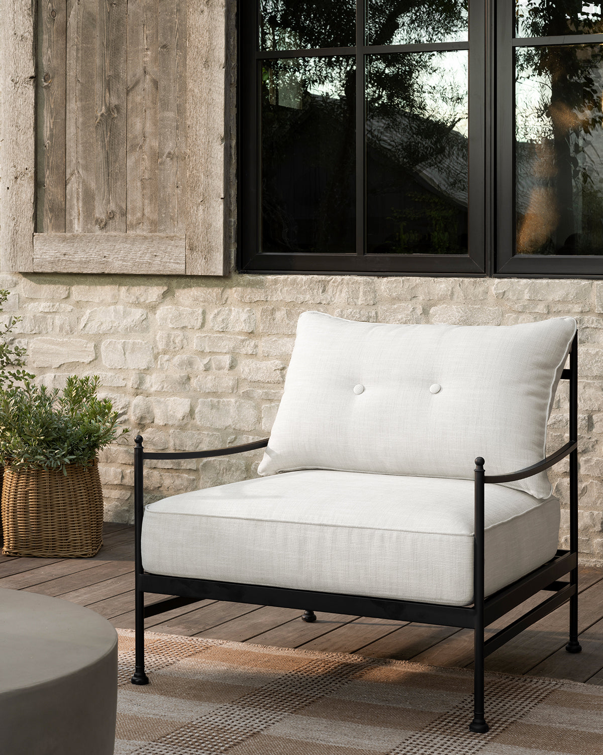 The Pomeroy Outdoor Lounge Chair with white performance cushions and a black powder-coated steel frame sits on a checkered rug by a basketed plant, in front of a stone wall and large window with wooden shutter by McGee & Co.