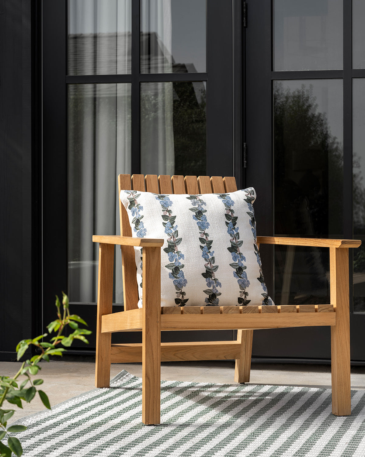 The Gilda Outdoor Lounge Chair, crafted from teak wood with a white cushion featuring blue floral patterns, sits on a striped rug before black-framed glass doors. A leafy plant appears in the foreground, by McGee & Co.