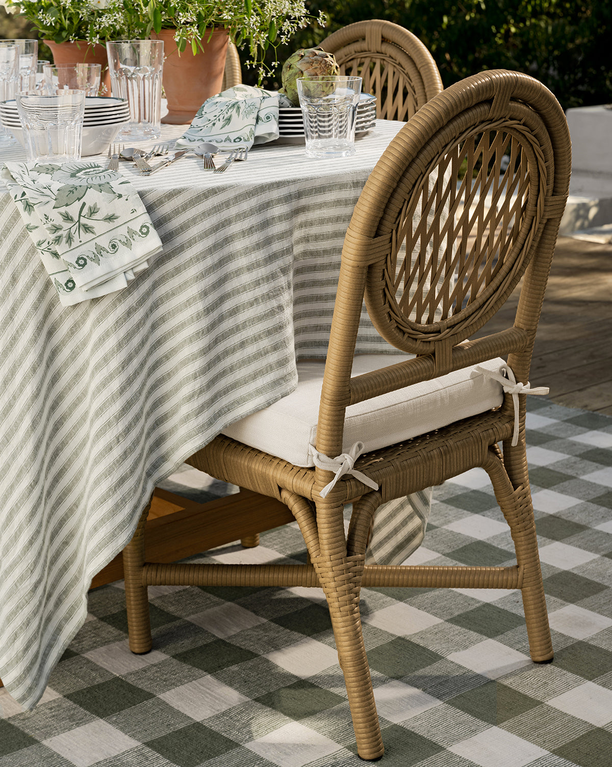 Two Marisol Indoor/Outdoor Dining Chairs with cushions sit at a table set with a striped tablecloth, glassware, plates, and patterned napkins on a checkered rug surrounded by greenery, by McGee & Co. Weather-resistant and handwoven wicker design.
