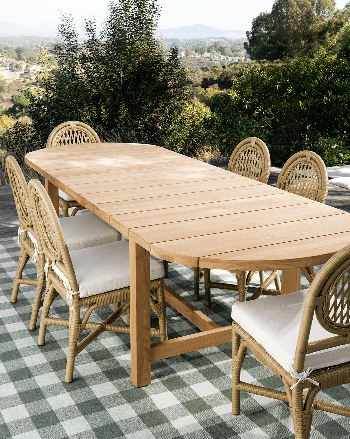 The Linwood Teak Extension Dining Table with eight wicker chairs sits on a green and white checkered rug on the patio, surrounded by lush trees and distant hills, by McGee & Co.