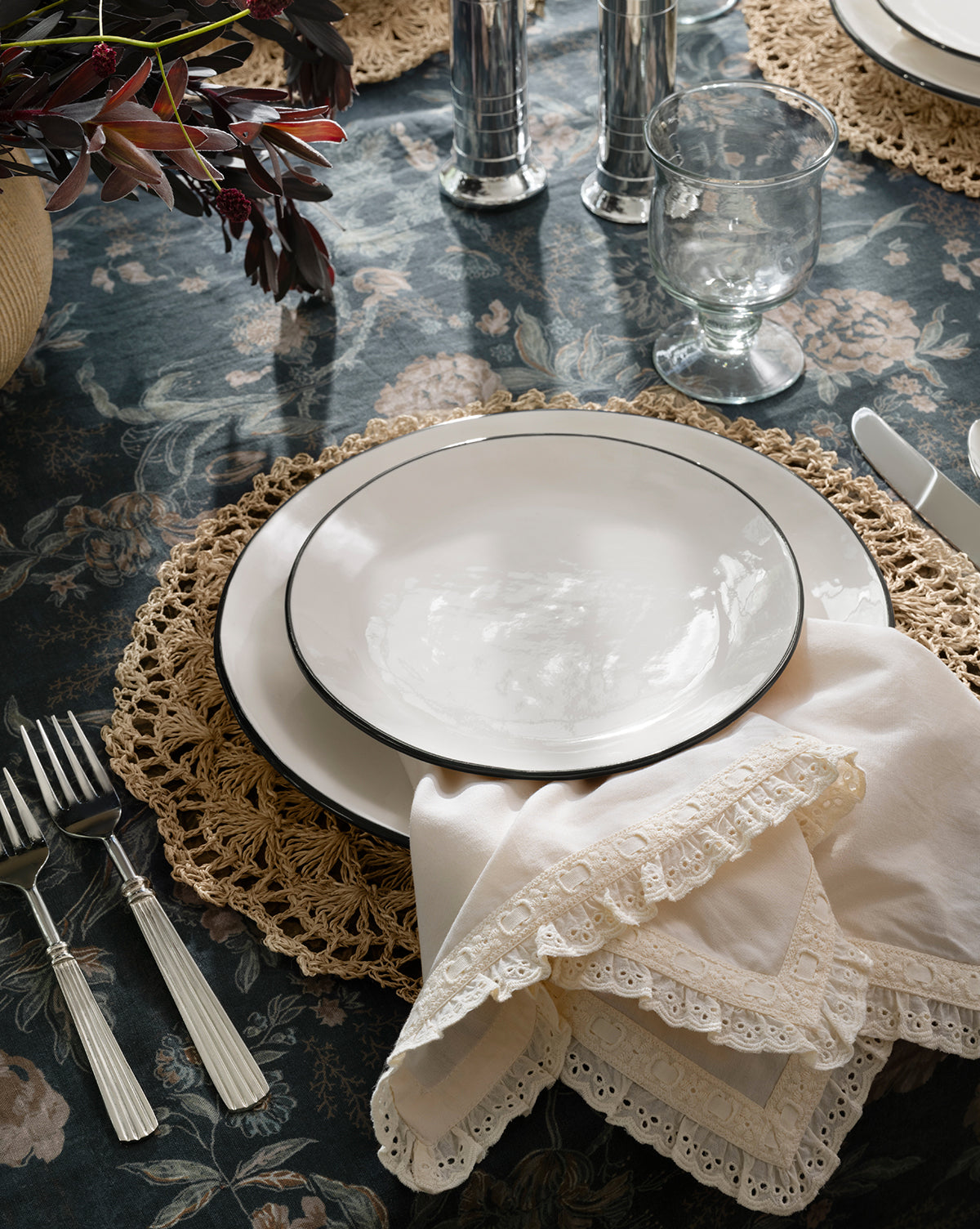 A place setting on a floral tablecloth features a white plate on a woven placemat, silver cutlery, a clear water glass, Lace Trim Napkins (Set of 4), and a vase with dark foliage in the corner, by McGee & Co.