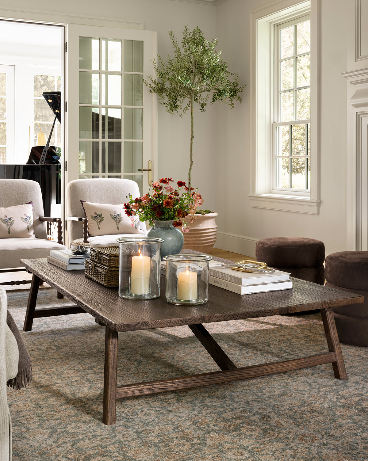A cozy living room features a wooden coffee table with candles, books, and flowers. Neutral sofas, poufs, a potted tree, and the Endicott Hand-Tufted Wool Rug enhance the bright space filled with natural light from large windows by McGee & Co.