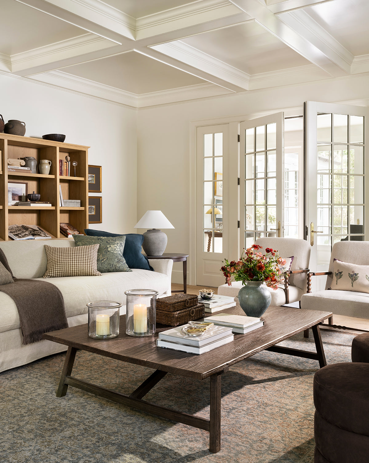 A cozy living room features a beige sofa, cushioned chairs, and the Lockwood Coffee Table. Shelves display decor, French doors fill the space with natural light, and the table holds candles, books, and a vase of flowers by McGee & Co.