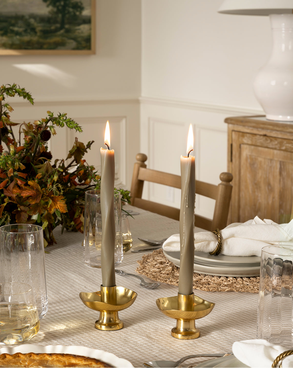 Two candles sit in the Cosima Taper Holder on a table, its antique brass design adding an elegant decorative touch to the scene, by McGee & Co.