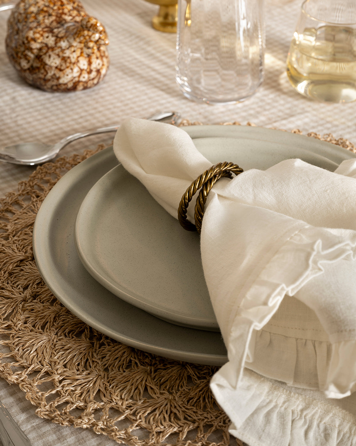 A close-up of a table setting features a white cloth napkin in a gold ring on the Aiden Soft Gray Dinner Plate, layered over beige plates on a woven placemat. A spoon, patterned glass, wine glass, and decor complete the elegant look, by McGee & Co.
