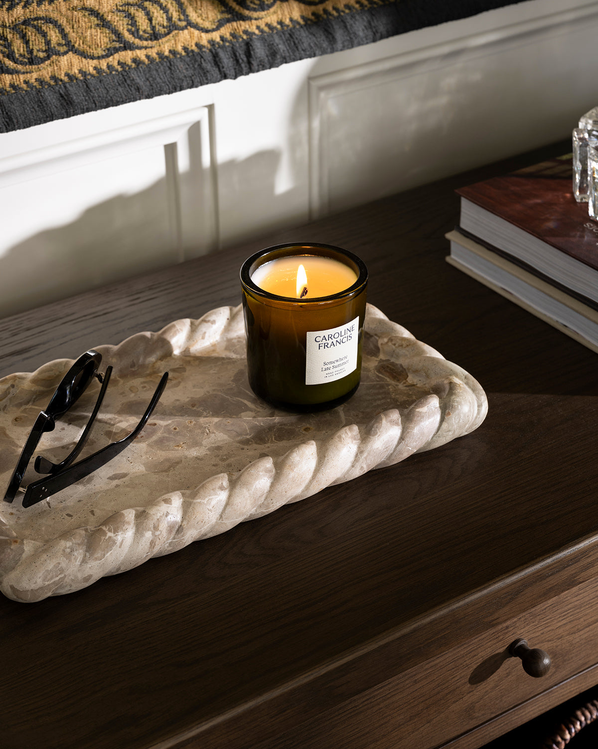 A lit scented candle in a glass jar sits on the Cleves Marble Tray next to eyeglasses on a dark wooden table, with books and a patterned bench adding cozy ambiance in the background, by McGee & Co.