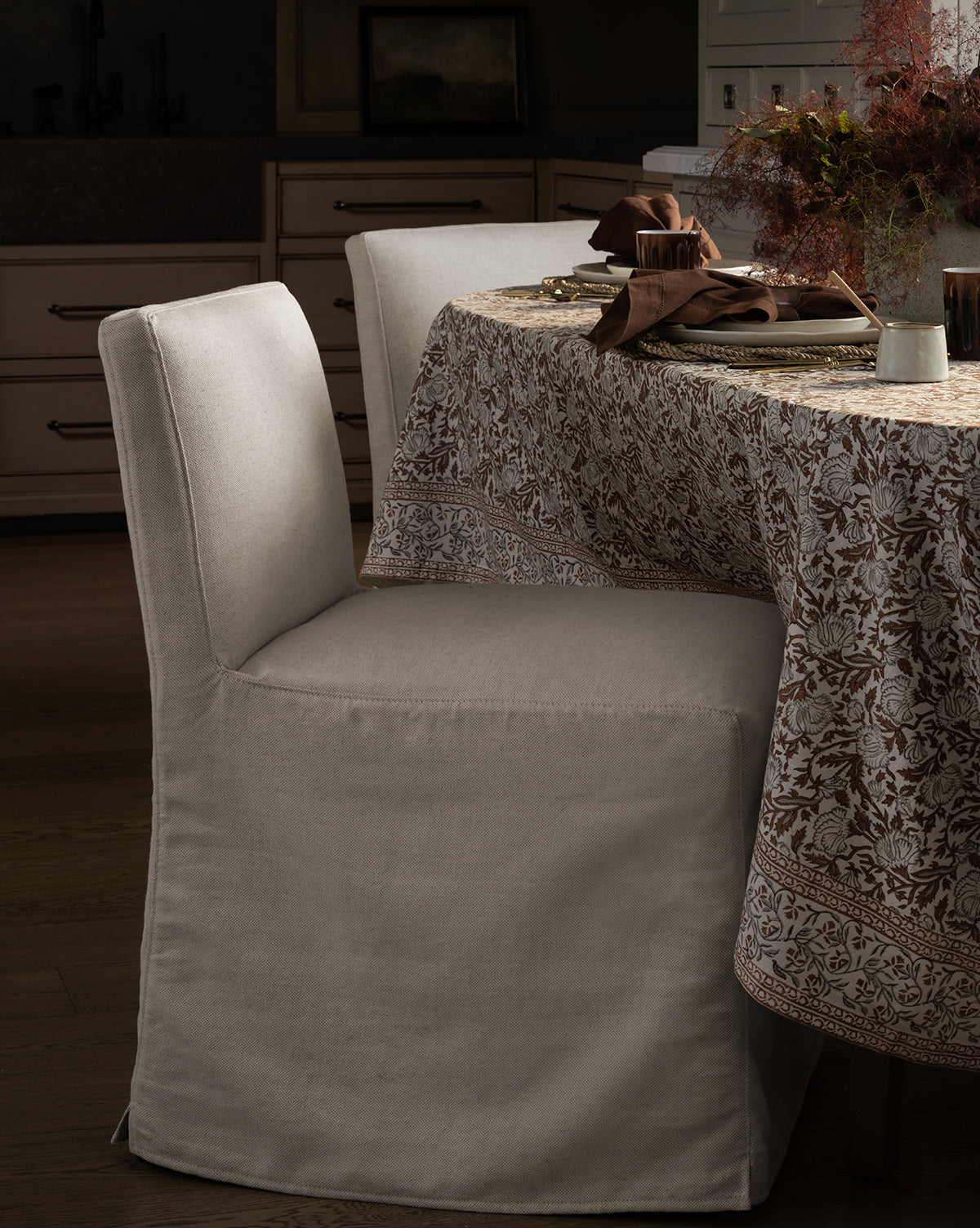 An Olivier Slipcover Dining Chair by Rowe Fine Furniture, Inc. sits next to a round table with a floral-patterned tablecloth, set with plates, cutlery, brown napkins, and a centerpiece of dried flowers, McGee & Co.