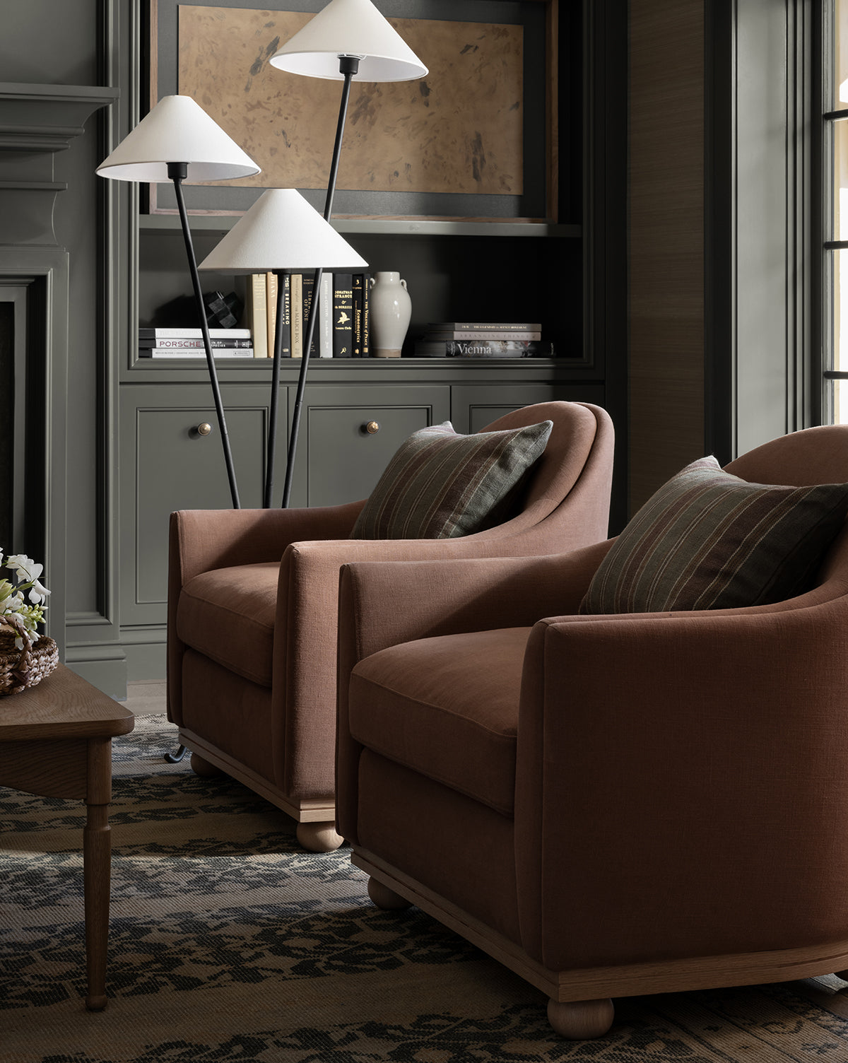 Two Bunnell Lounge Chairs (Ready to Ship) with striped pillows are placed by a wooden coffee table in a cozy room. Behind them, a dark grey built-in bookshelf holds books, vases, and three white-shaded floor lamps, by McGee & Co.