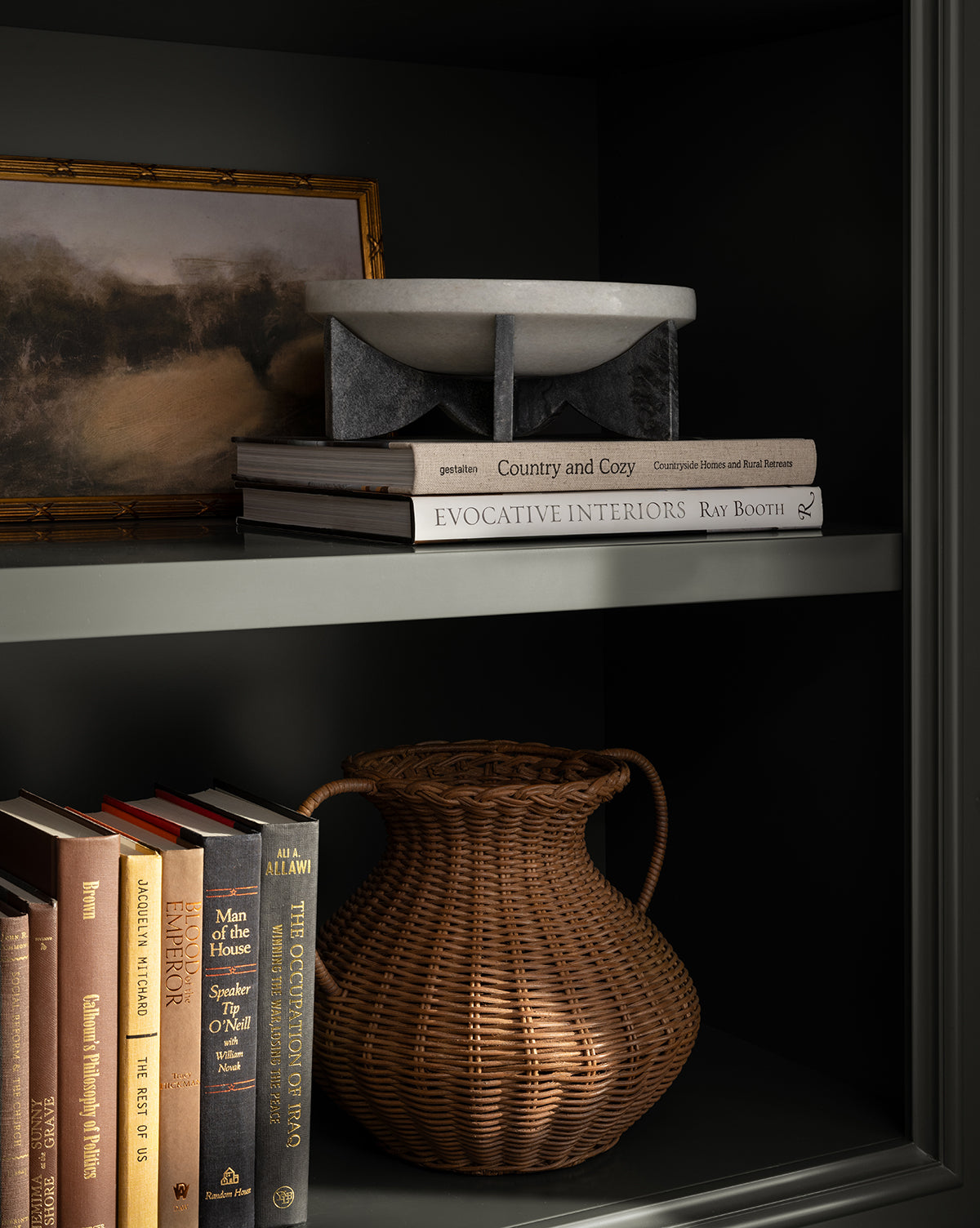 A bookshelf displays a framed landscape painting, the Mahoney Bowl with its sculptural white marble design, stacked books including Country and Cozy and Evocative Interiors, plus a large woven wicker vase on the lower shelf beside standing books by McGee & Co.