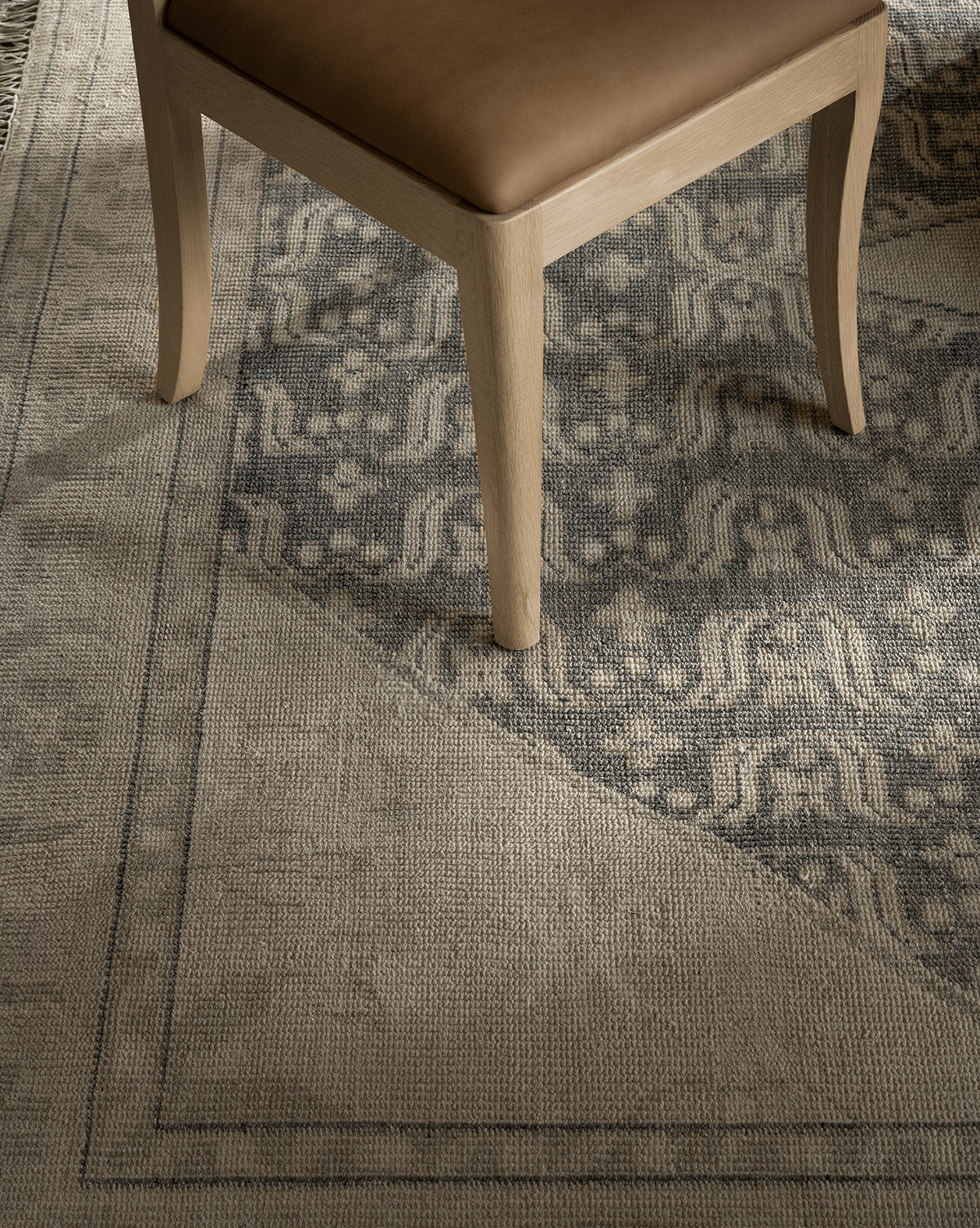 A close-up of a wooden chair with a tan seat cushion sits on the Elin Hand-Knotted Wool Rug, which displays geometric and floral motifs in muted gray and beige, showcasing its artisan craftsmanship, by McGee & Co.