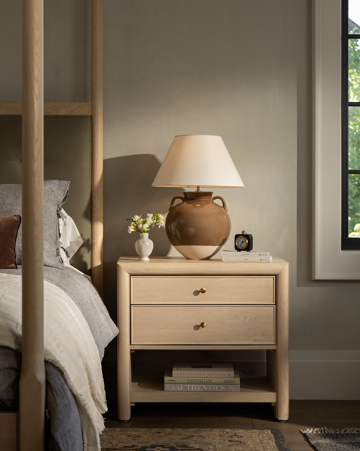 A Tellerbeck Nightstand sits by the bed, featuring two drawers and topped with a brown lamp, white vase of flowers, and black alarm clock—ideal for stylish bedroom organization as sunlight filters in by McGee & Co.