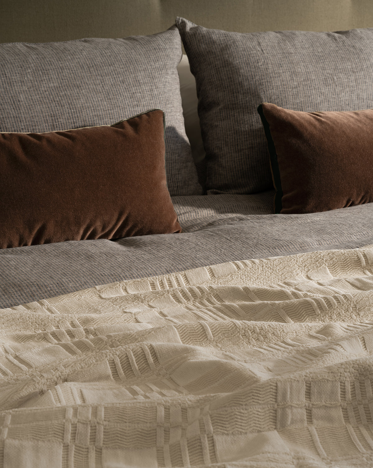 The inviting cream bedding ensemble is completed by the Makerspalm Chamberlain Coverlet, styled with textured gray pillows, brown velvet rectangular cushions, a gray patterned duvet, and a cream woven blanket at the foot—McGee & Co.