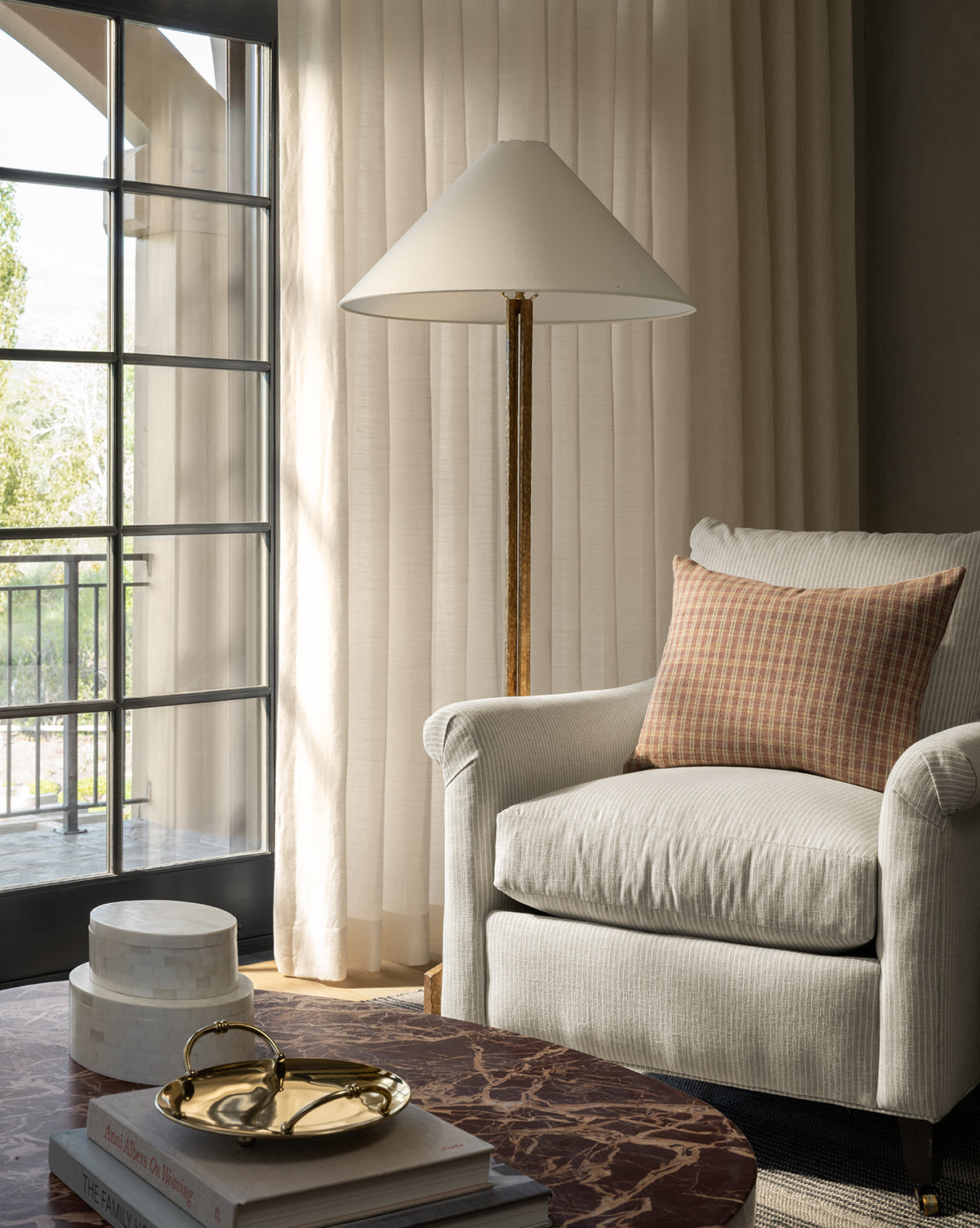 A cozy living room corner features the Reedley Lounge Chair by Rowe Fine Furniture, Inc., a brown checkered pillow, gold floor lamp, sheer curtains, and a marble coffee table with books and decorative boxes near a large window—finished with an accent from McGee & Co.