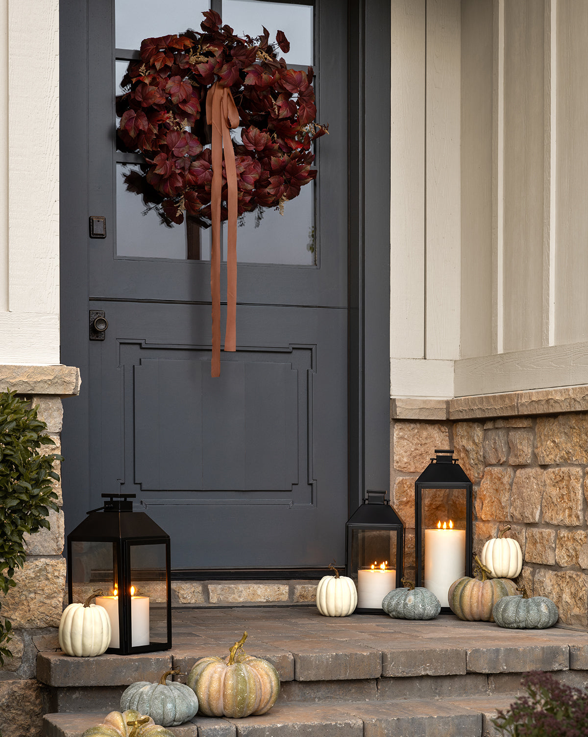 A dark blue door features a Faux Autumn Grapevine Wreath with a brown ribbon, complemented by seasonal decor—white and green pumpkins, faux greenery, and black lanterns with candles—set against a charming stone exterior.