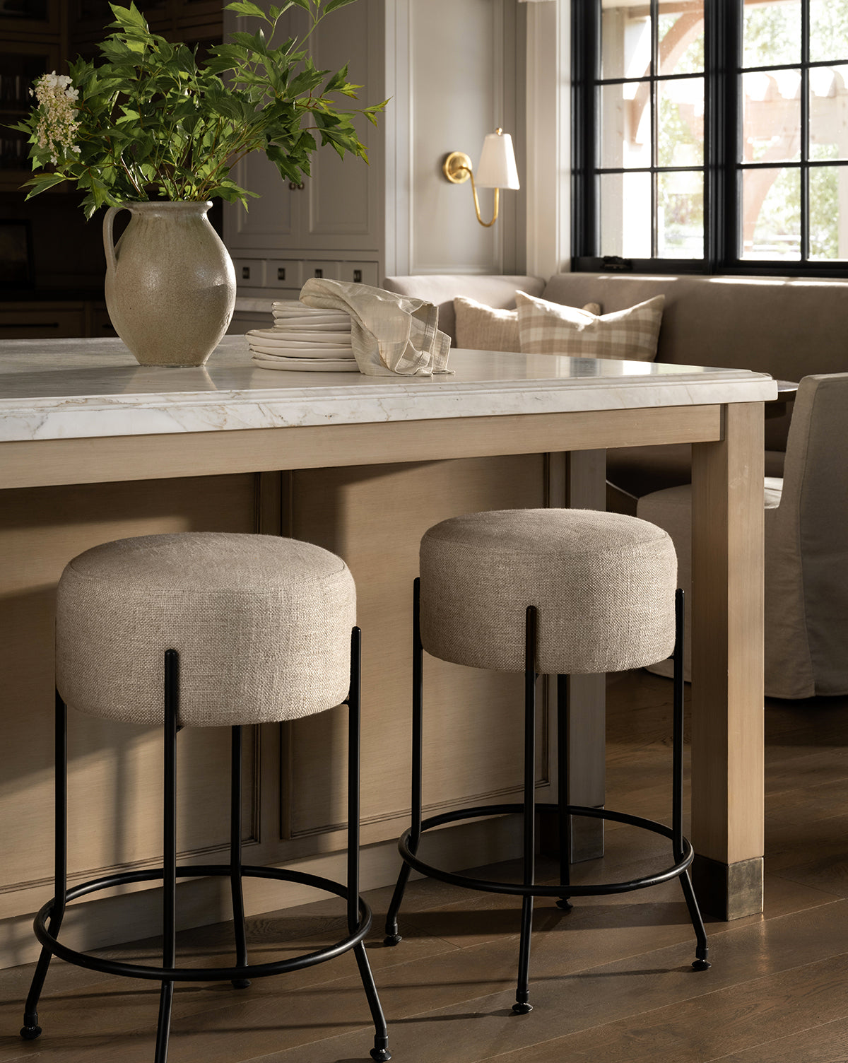 Certainly! Here’s your revised sentence with McGee & Co. blended at the end: A modern kitchen showcases a marble countertop island with a beige vase of greenery, stacked white plates, and two Makerspalm Evander Counter Stools—industrial-inspired with black metal legs—plus a window seat in the background, all styled by McGee & Co.