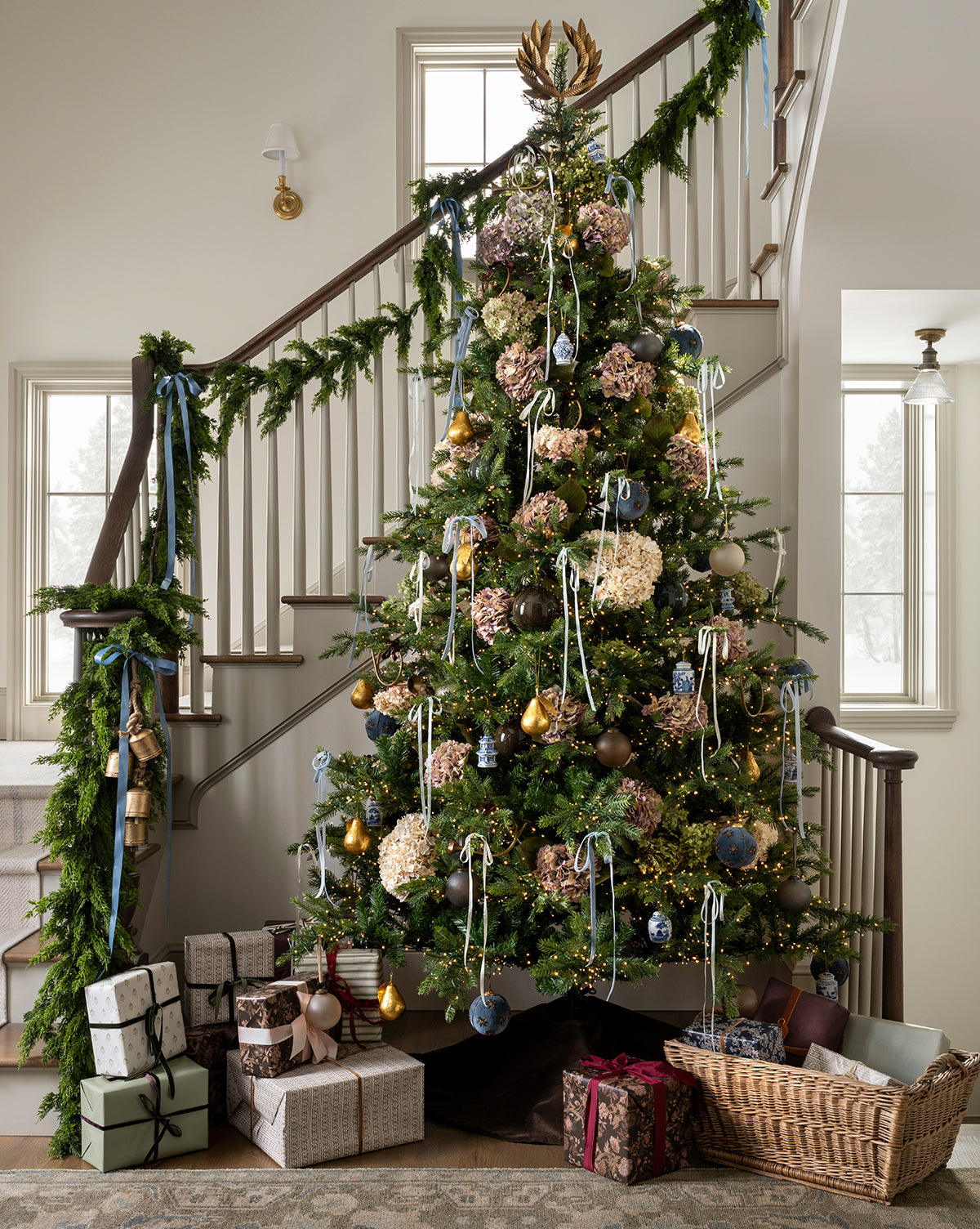 A decorated Christmas tree stands by a staircase draped with the 5' Cedar Faux Garland. Wrapped gifts and a basket are arranged on the floor around the tree in a bright, cozy room, by McGee & Co.