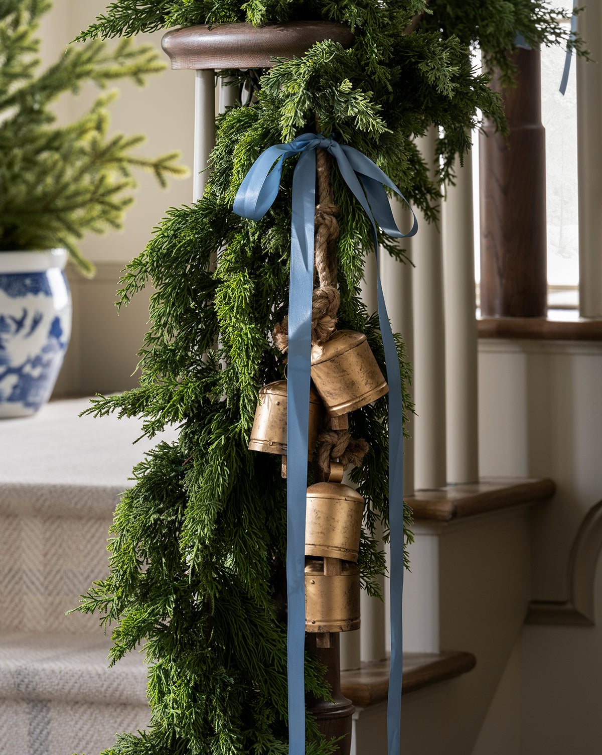 A staircase railing decorated with a 5' Cedar Faux Garland, brass bells tied with a blue ribbon, and a potted plant in a blue and white patterned pot in the background, by McGee & Co.