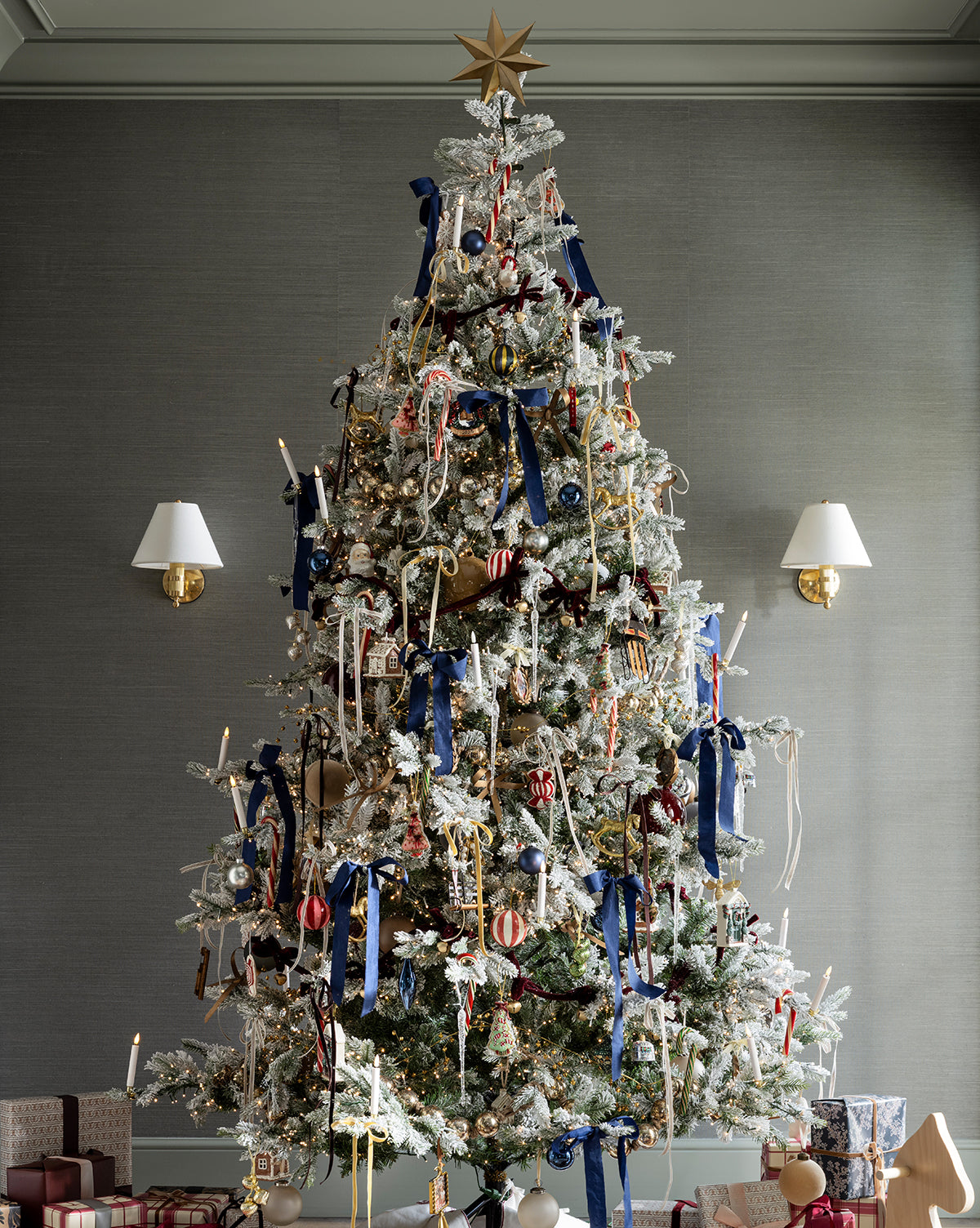 A tall holiday tree stands in a stylish room with gray walls, adorned with blue ribbons, white lights, ornaments, and candy canes. At the top shines the Etoile Tree Topper. Wrapped presents sit at the base, flanked by brass wall sconces, by McGee & Co.