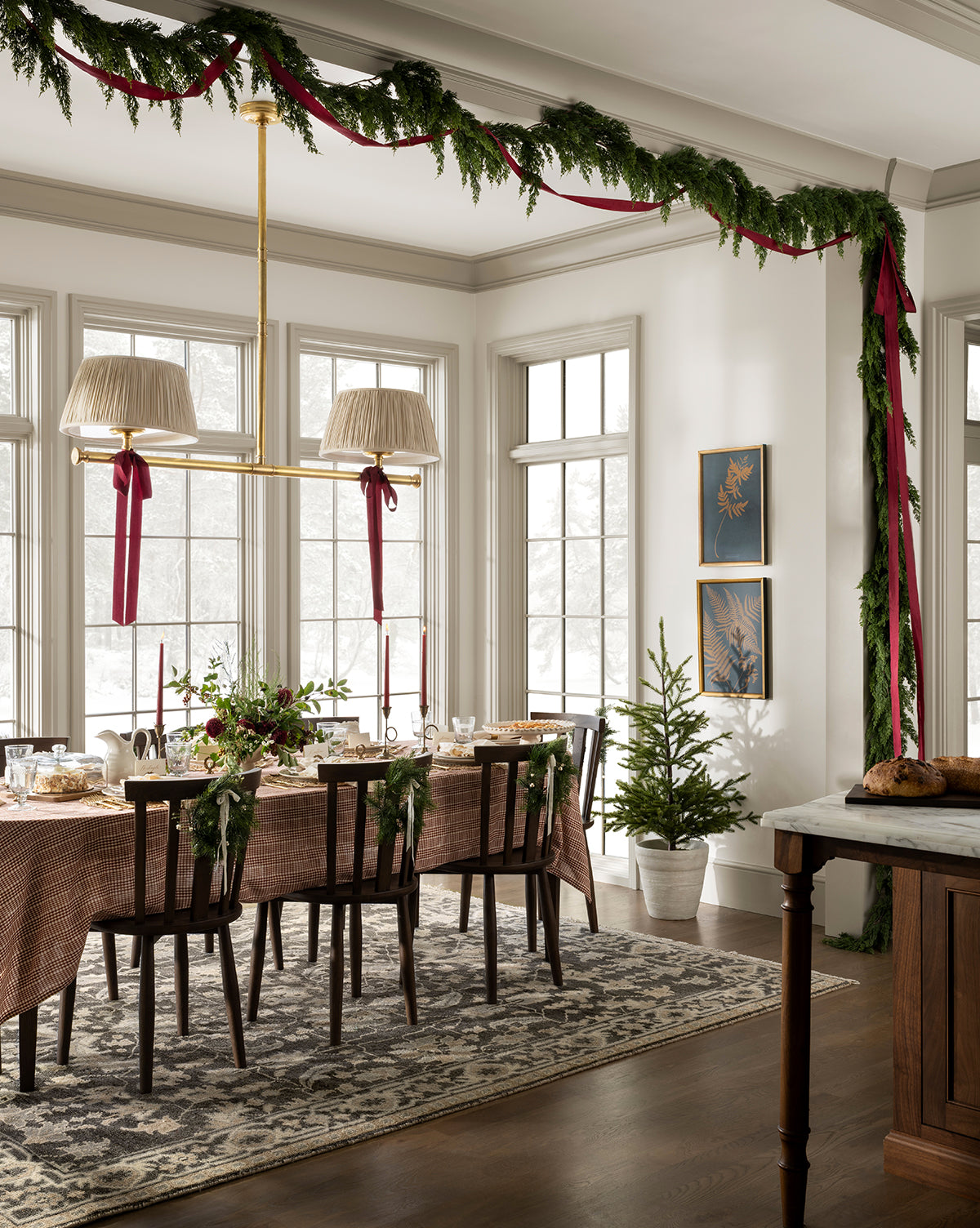 A dining room features a 6' Cedar Pre-Lit Faux Garland, red ribbons, a festive table, two small potted Christmas trees, and large windows streaming natural light—perfect for holiday home decor, by McGee & Co.