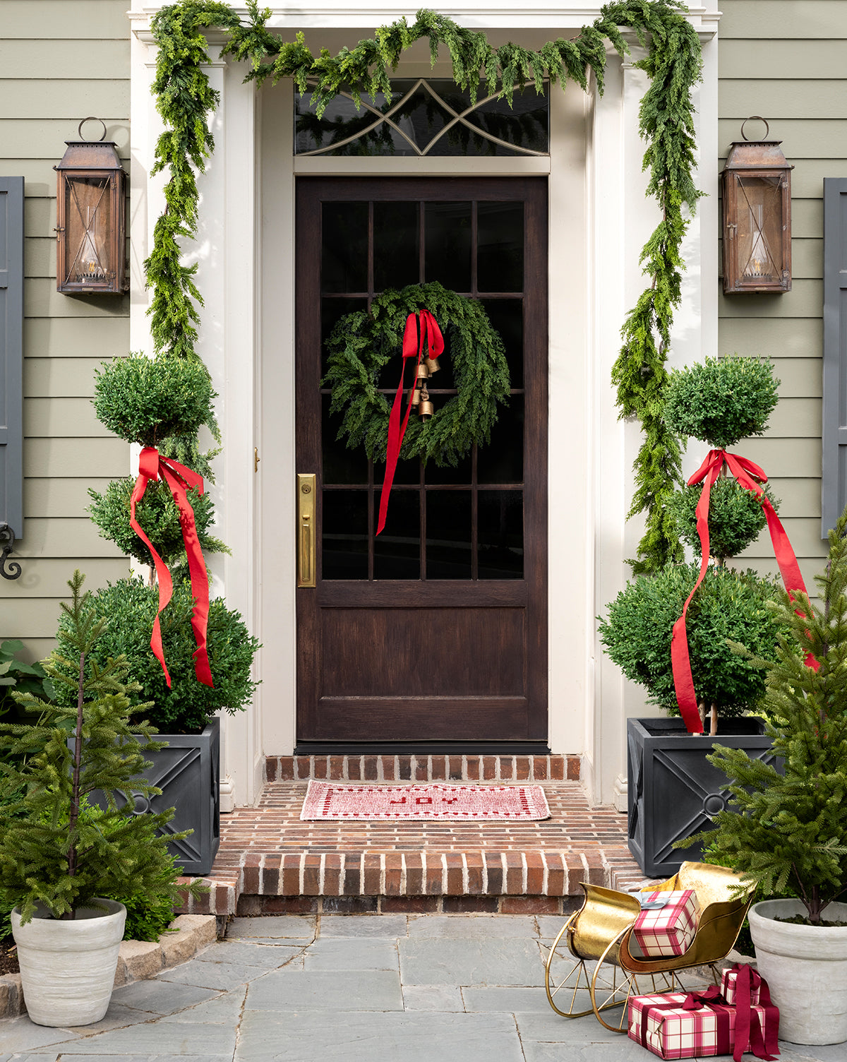 A front door decorated for Christmas with a wreath, red ribbon, and a 5' Cedar Faux Garland draped around the frame, flanked by potted topiary trees with red bows, small evergreens, and wrapped gifts on the doorstep, by McGee & Co.