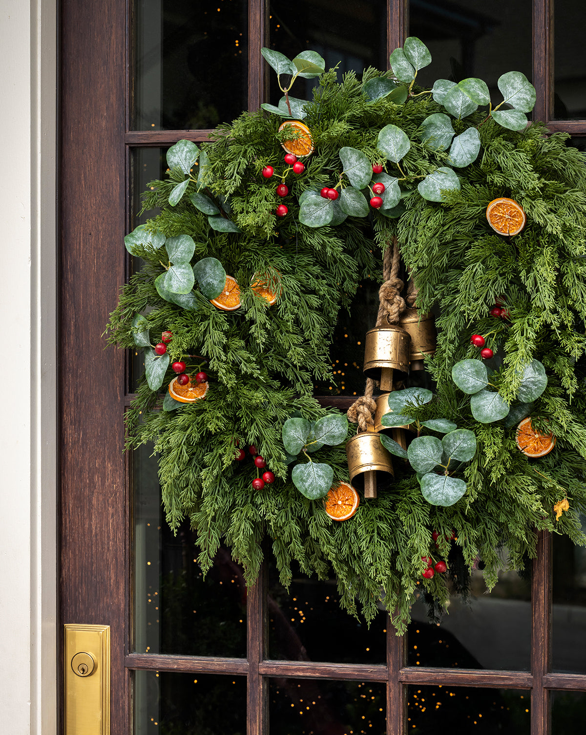 The 30" Orange & Eucalyptus Faux Wreath, featuring evergreen branches, eucalyptus leaves, red berries, dried orange slices, and gold bells, hangs on a dark wooden door with a brass handle, by McGee & Co.