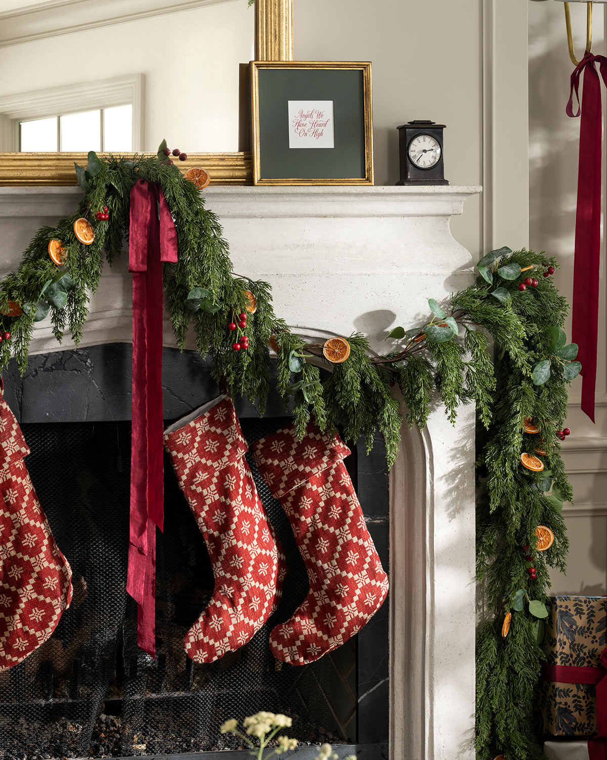 A fireplace is adorned with a 6' Orange & Eucalyptus Faux Garland, featuring lush greenery and orange accents. Three red patterned Christmas stockings hang from the mantel, paired with cozy holiday decor like a framed sign and a clock, by McGee & Co.