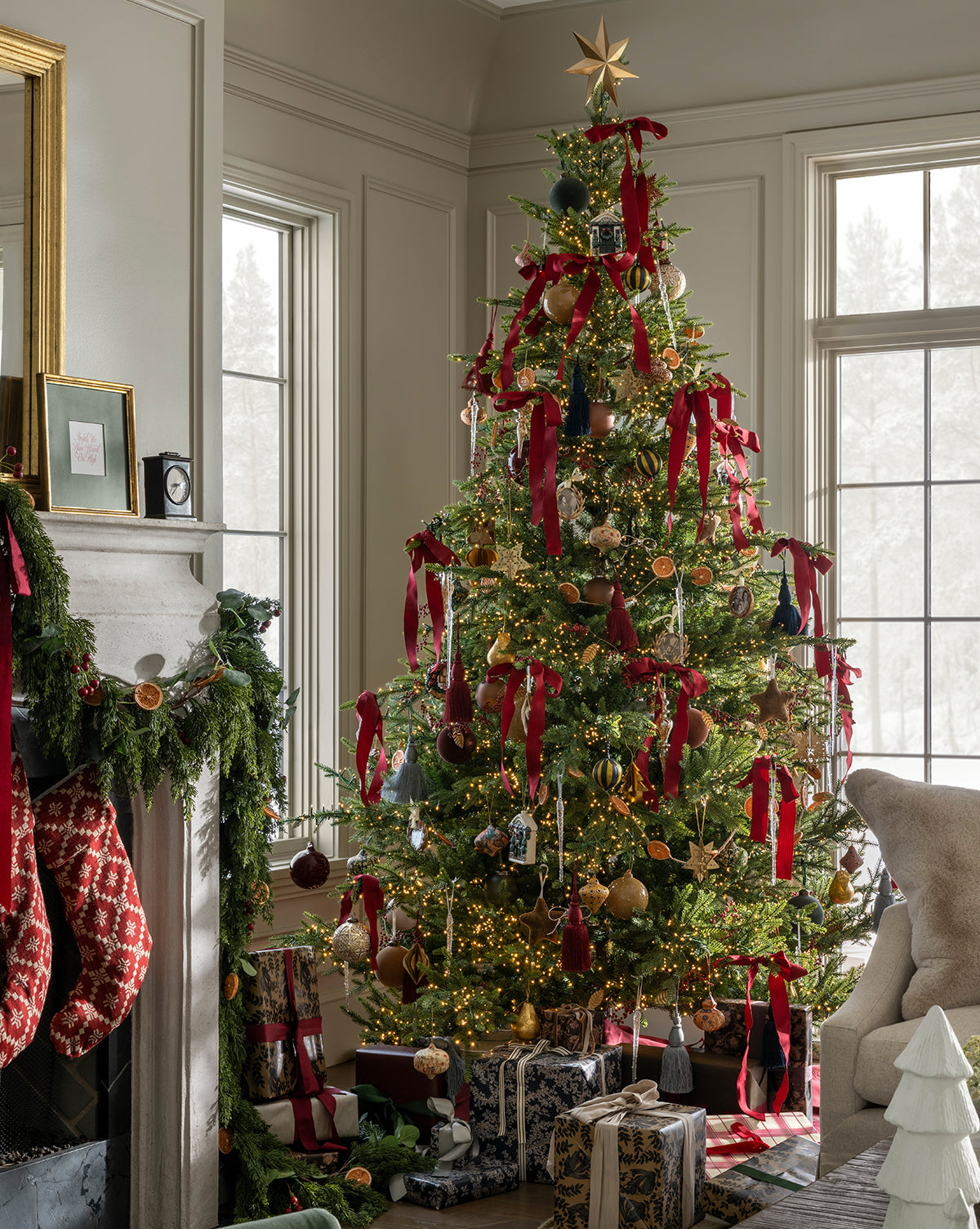 A holiday tree trimmed with red ribbons, ornaments, and lights is topped with the Etoile Tree Topper. Wrapped gifts rest below, while festive stockings hang from a fireplace draped in garland nearby, by McGee & Co.