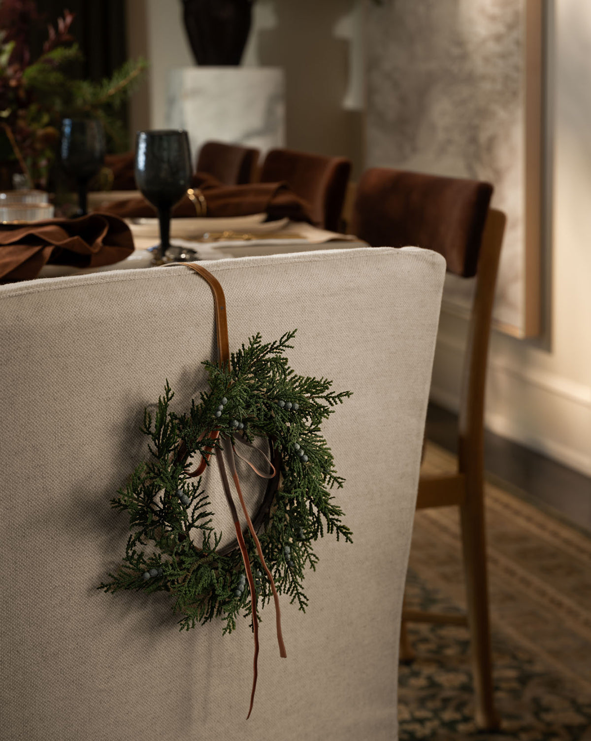 An 8" Blueberry & Juniper Faux Wreath with ribbons hangs on the back of a beige dining chair at a table set with dark glassware and brown napkins, creating an elegant, festive holiday atmosphere, by McGee & Co.