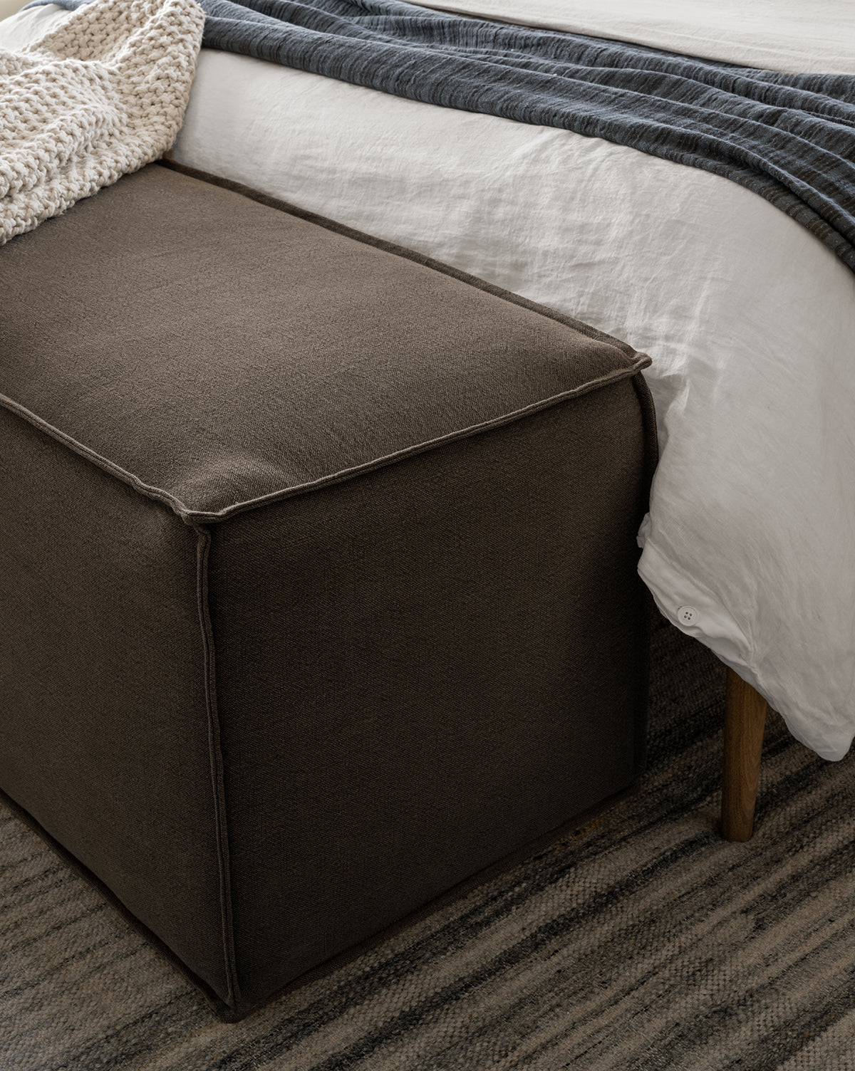 A dark brown Johannes Bench is placed at the foot of a bed with white bedding, a gray throw, and a textured rug, next to a chunky knit blanket by McGee & Co.