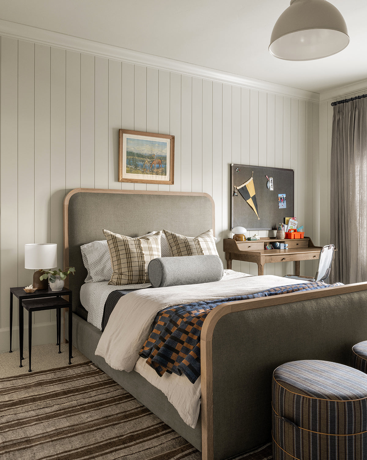 A cozy bedroom features the Community Ainsworth Bed, plaid pillows, striped bedding, a side table lamp, a striped rug, a desk with bulletin board, sheer window curtains, and art hanging above the bed—McGee & Co.