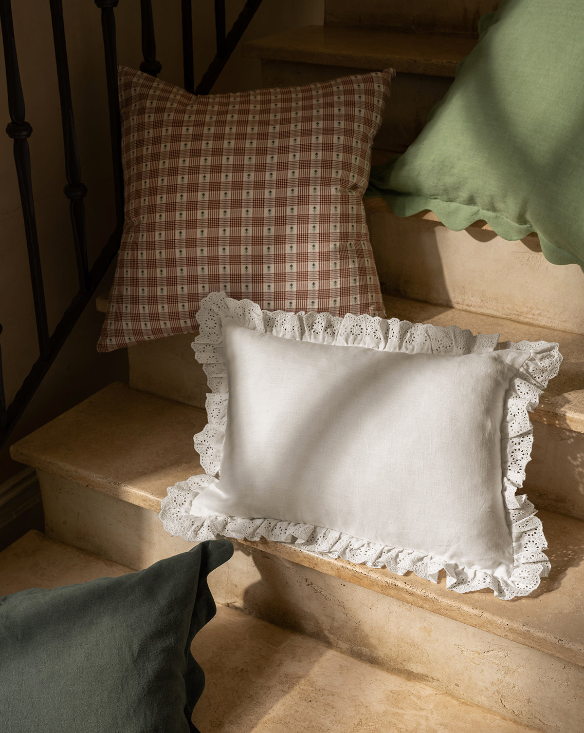 Three decorative pillows rest on stone steps: a green pillow with a wavy edge, a brown checkered cushion, and the McGee & Co. x Loeffler Randall Delilah Broderie Trim Pillow Cover in white linen. Sunlight casts soft shadows across the scene by McGee & Co.