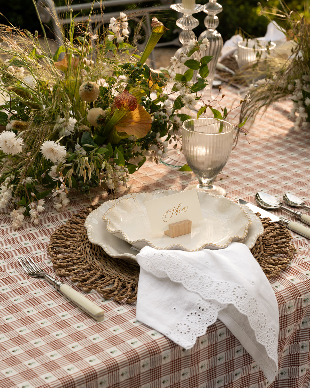 A rustic outdoor table set features a checked tablecloth, woven placemat, scalloped plate, McGee & Co. x Loeffler Randall Delilah Broderie Napkins (Set of 4), glassware, silver cutlery, a name card, and wildflowers in natural light by McGee & Co.