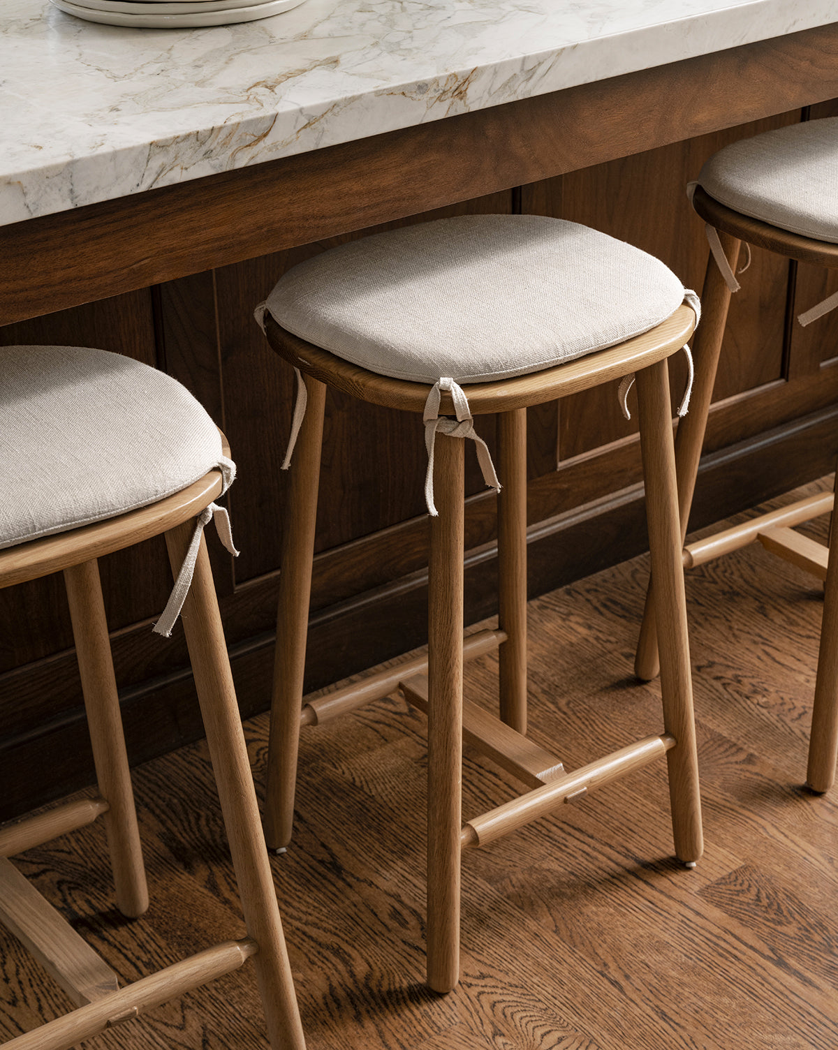 Three Ralph Oak Counter Stools with linen cushions and ribbon ties sit under a marble kitchen counter with dark wood paneling, all on a wooden floor by McGee & Co.