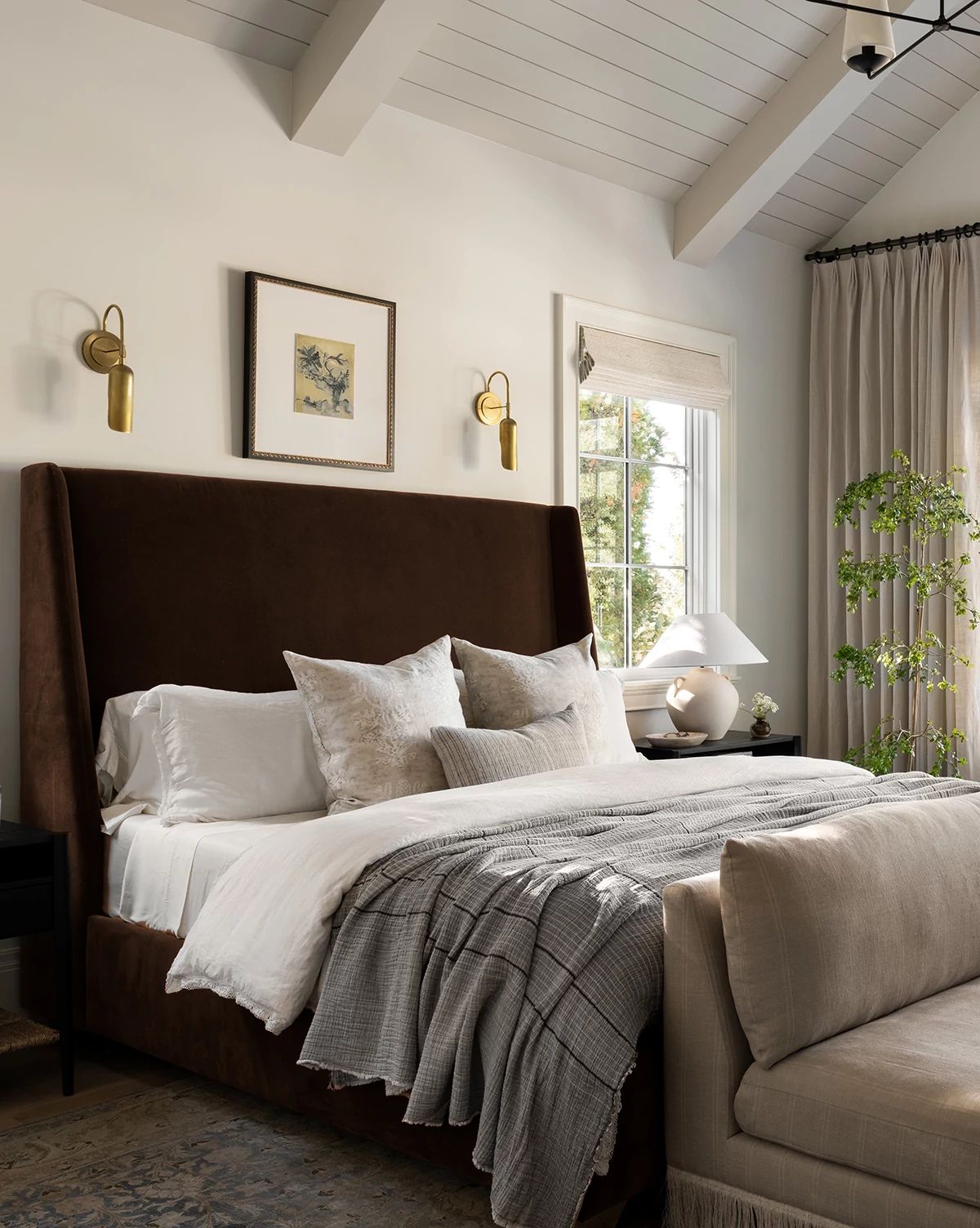 A bedroom featuring the Walt Bed Tall Headboard in dark brown with solid oak legs, white bedding, neutral pillows, gray blanket, two wall sconces, framed art, window curtains, and a leafy plant beside the bed by McGee & Co.