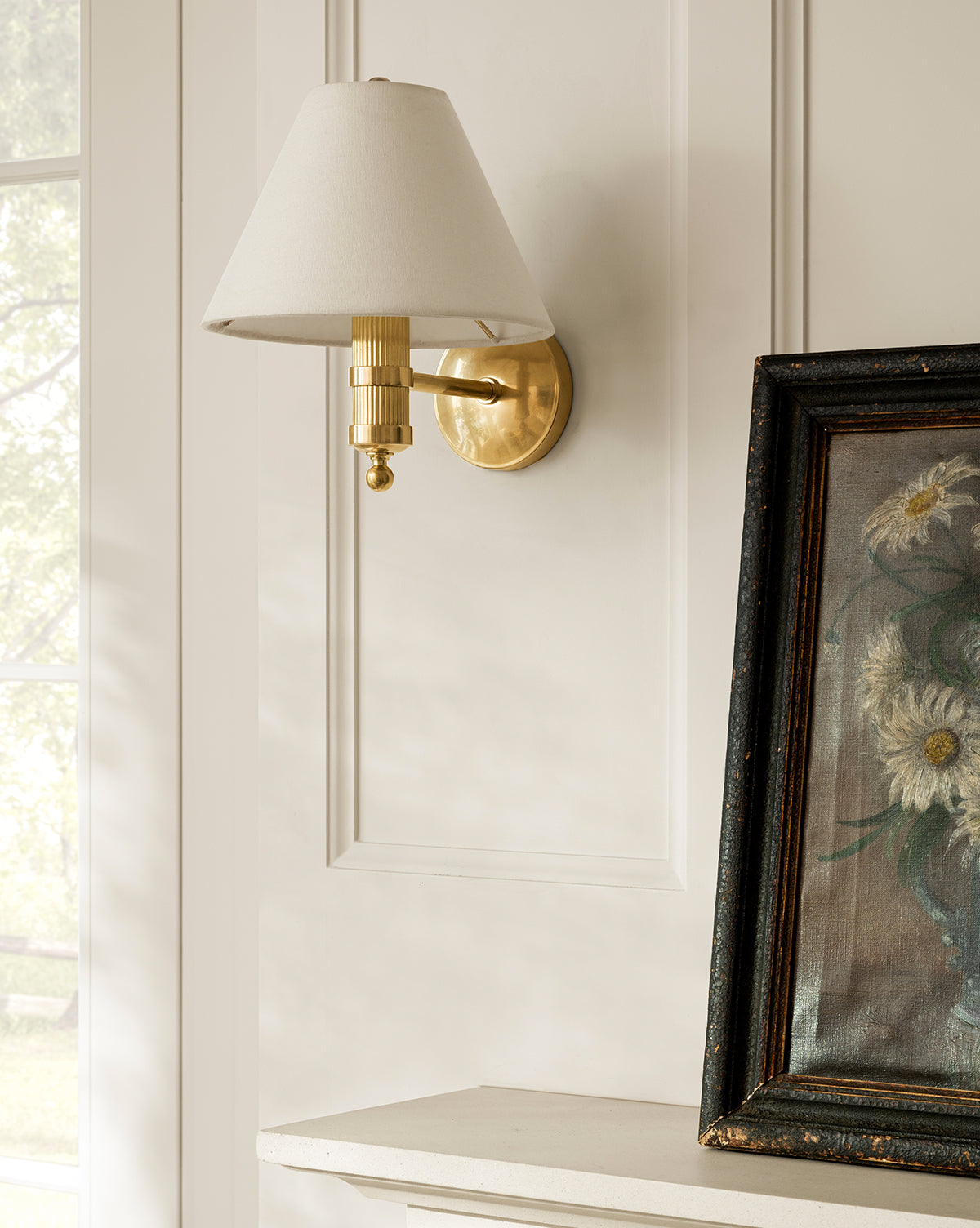 A Meredith (China) Ltd Townes Sconce with a linen shade is mounted on a white paneled wall above the mantel, beside a framed floral painting. Sunlight pours in from the window to the left, McGee & Co.