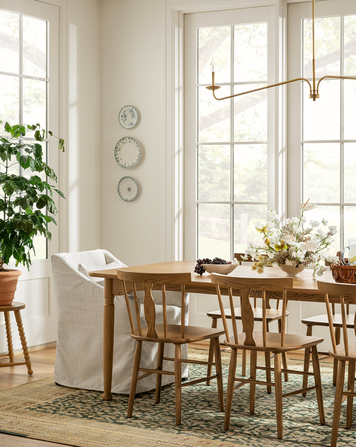 A bright dining space with large windows, a solid oak table, mismatched chairs including the Whittaker Chair by Yang Chen, with a potted plant, and floral centerpiece. Light fills the cozy room. McGee & Co.