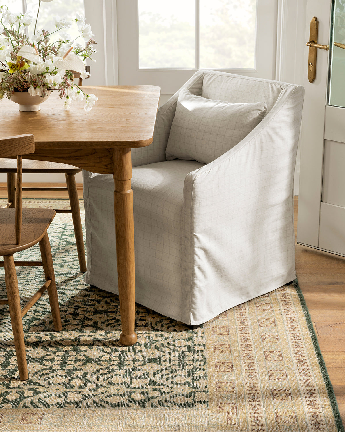 An Olivier Slipcover Dining Arm Chair by Rowe Fine Furniture, Inc. in light gray with a feather and down cushion and matching pillow sits at a wooden dining table beside another chair, atop a patterned rug, with floral accents and natural light nearby. McGee & Co.