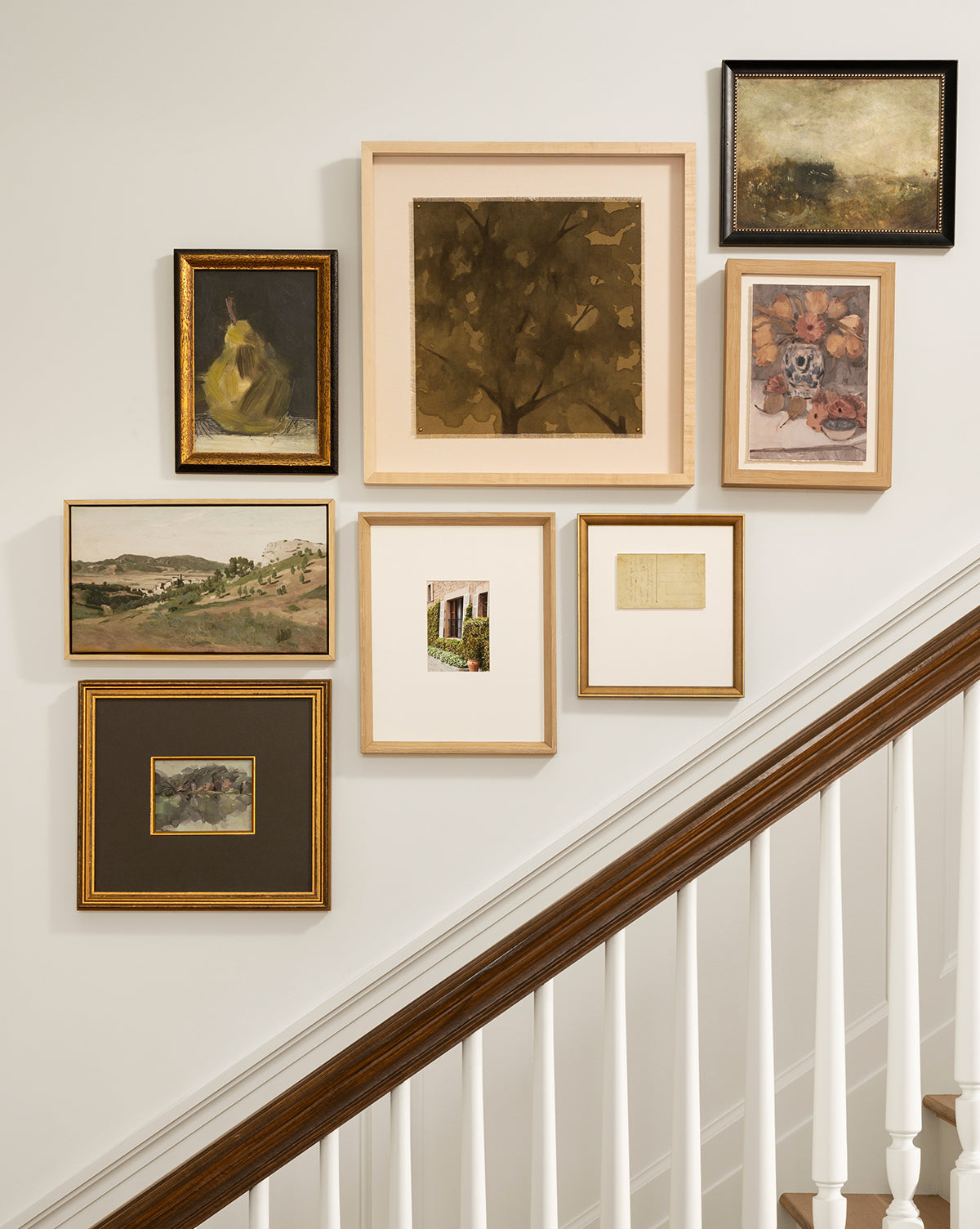 A gallery wall above a staircase features seven artworks, including the View of the Town canvas in an oak frame. The balanced cluster pops against a cream wall, with a white railing and wooden banister visible in the foreground, by McGee & Co.