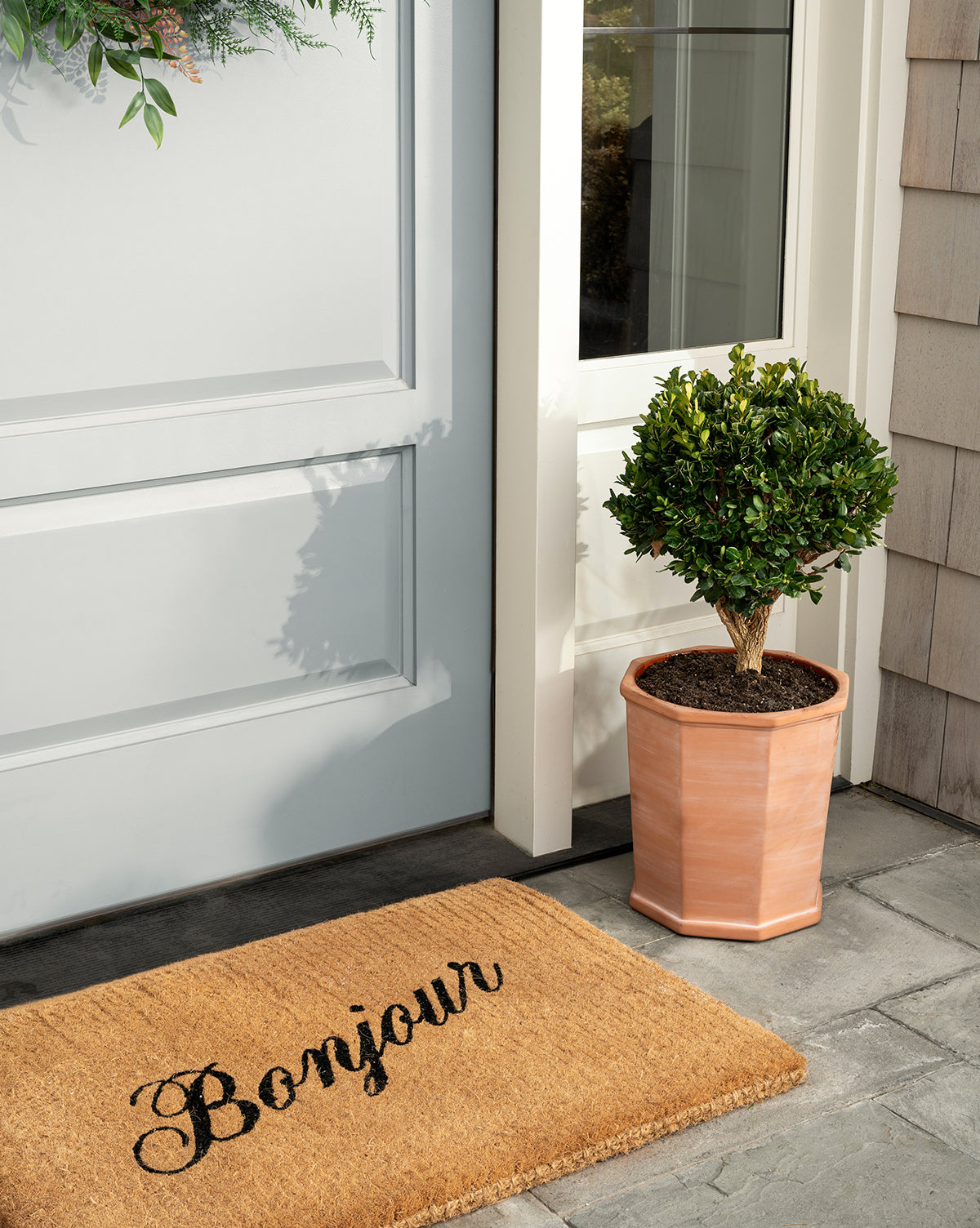 A handwoven coir Bonjour Doormat rests before a light blue door, bringing charm to your entryway, while a potted topiary in a light brown planter completes the welcoming scene, by McGee & Co.