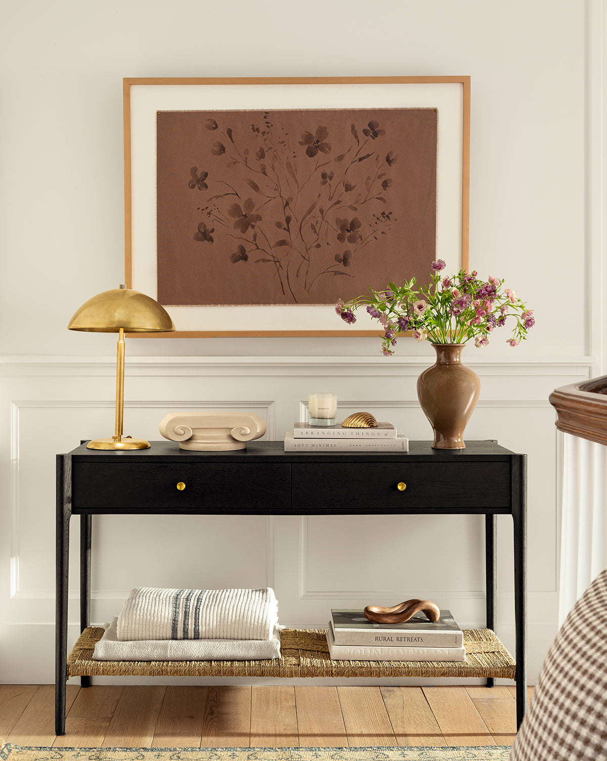 A stylish entryway features a black console table with gold knobs, decorated with a brass lamp, books, purple flowers in a vase, and the Consider the Wildflowers by Katie Mulder print in a maple frame above, plus folded blankets on the lower shelf, by McGee & Co.