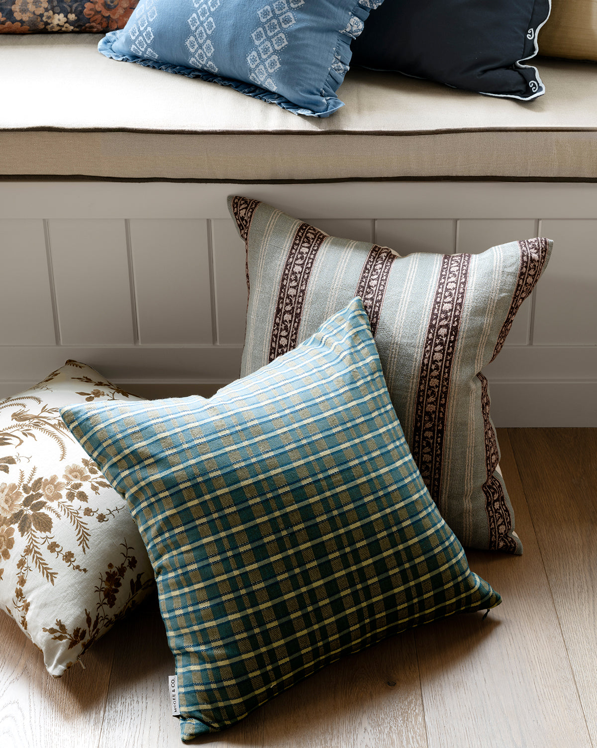 Three decorative pillows rest on a wooden floor: one with the Selwyn Plaid Pillow Cover in blue and green, one with brown and white stripes, and one with a brown floral print. Behind them, a cushioned bench with pillows adds timeless charm, by McGee & Co.