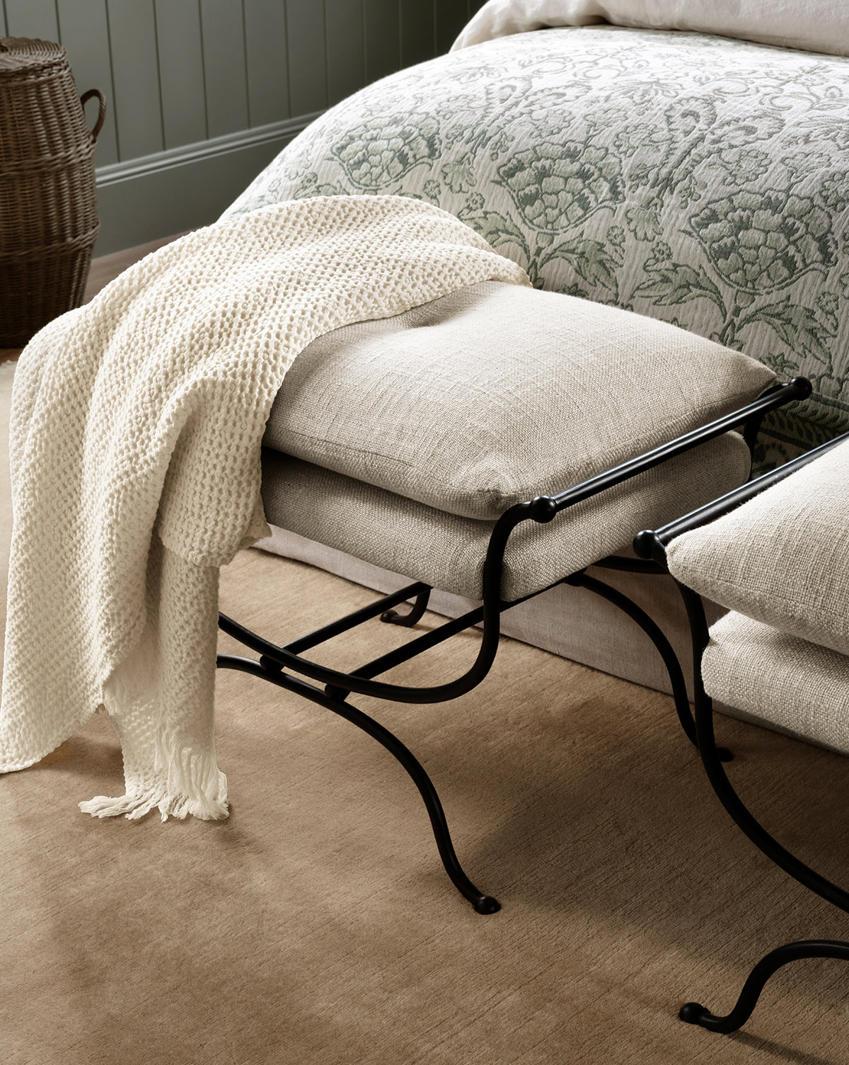 A cushioned bench with a black metal frame sits by a bed with a floral comforter. The cream Viola Throw, featuring a textured weave, is draped over the bench, and a wicker basket rests on the beige carpet in the background, by McGee & Co.