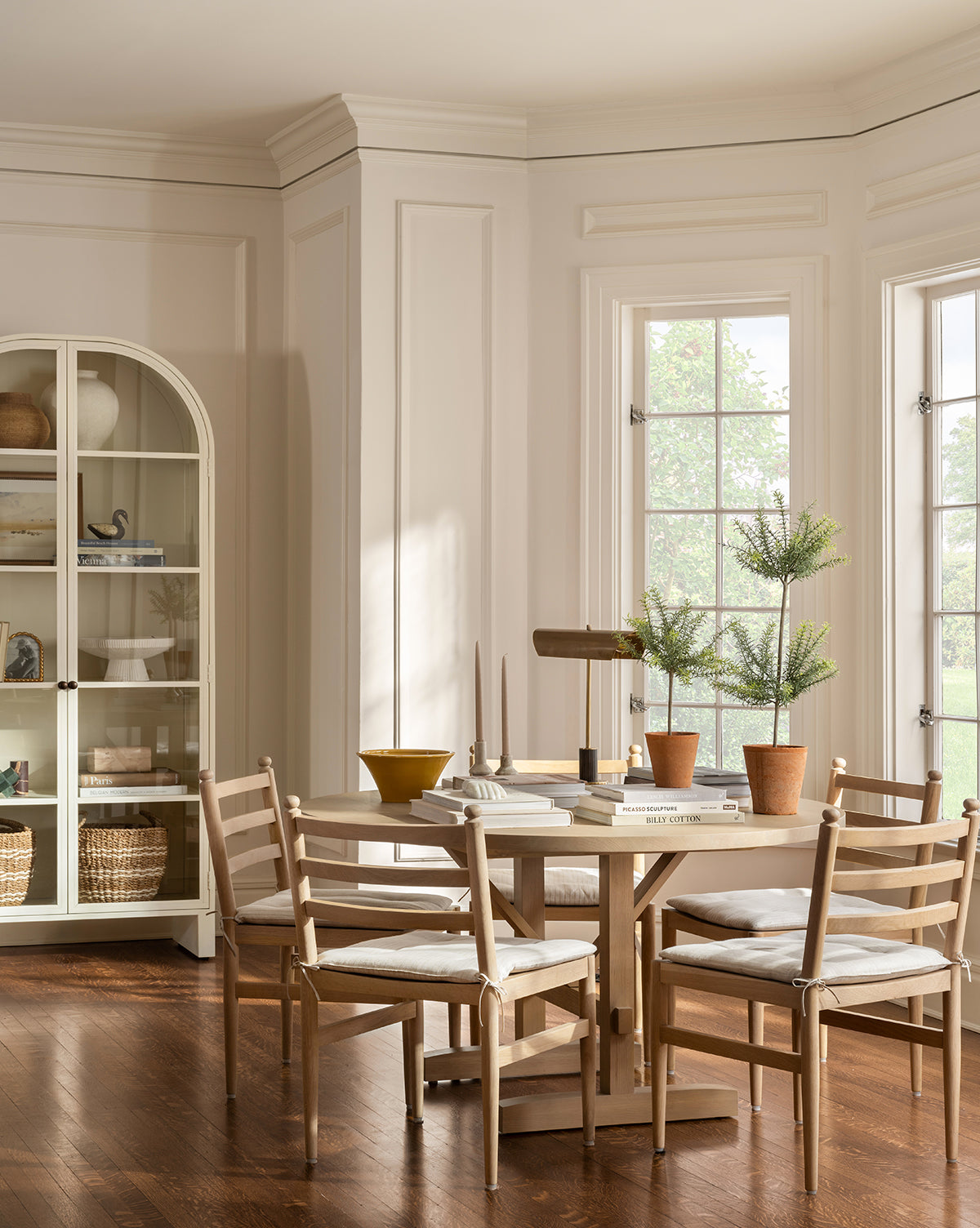 A bright dining room features the Giselle Round Table, surrounded by five cushioned wood chairs. Potted plants, books, and decor adorn the tabletop. Large windows illuminate the space, with a white cabinet visible in the background, by McGee & Co.