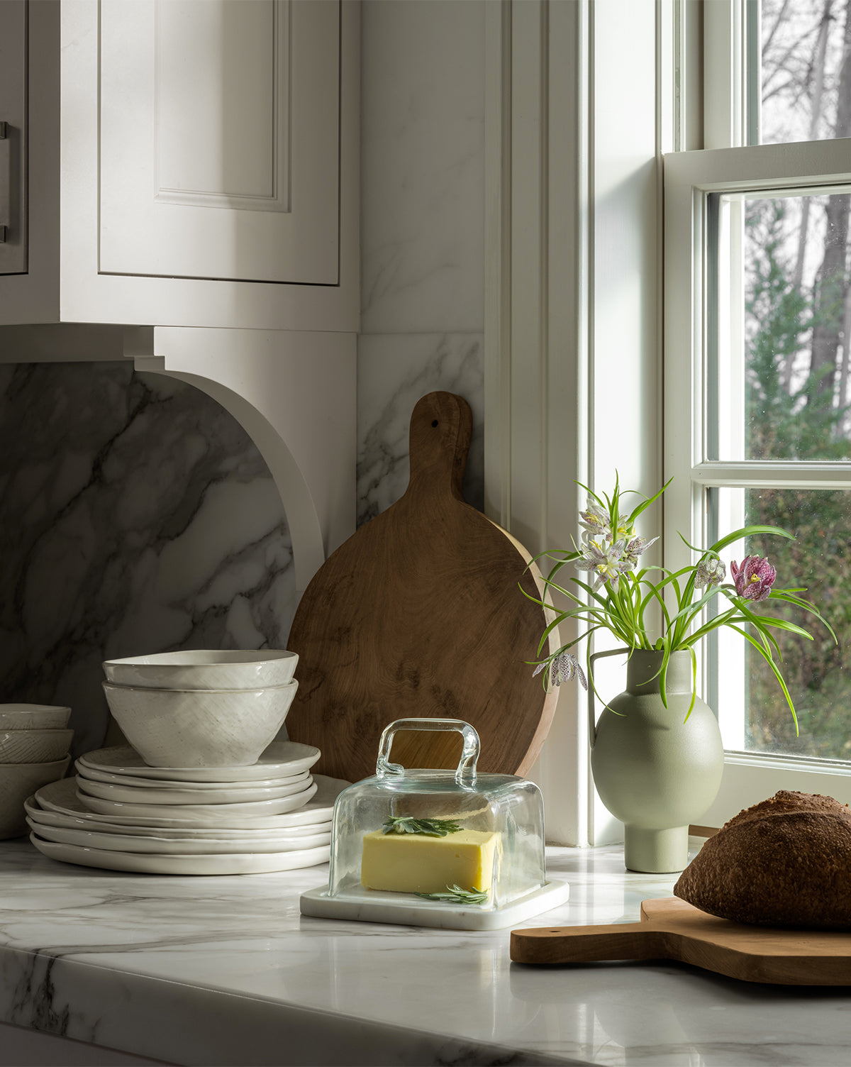 A marble kitchen countertop with stacked white dishes, a vase of flowers, and a loaf of bread on the Boylan Bread Board sits by a window in natural light, with butter under a glass cover nearby.