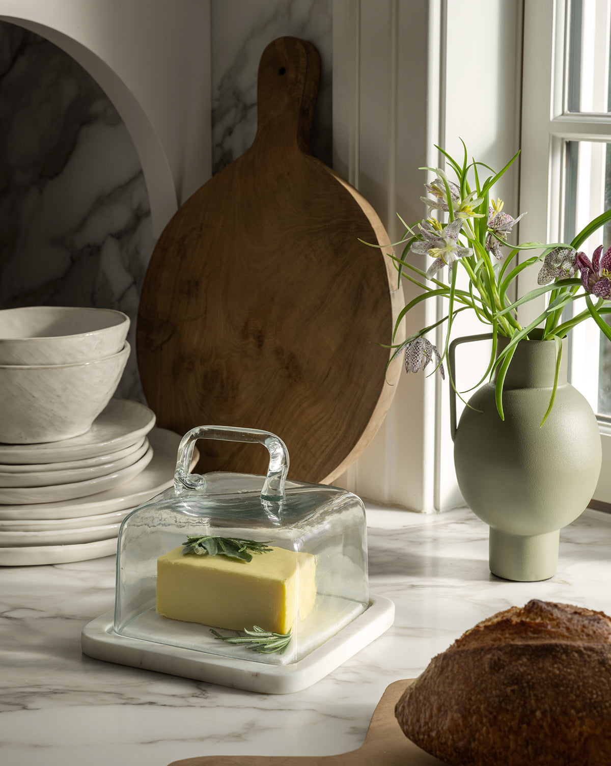 A stick of butter with herbs sits under a glass dish on a marble counter beside bread, stacked white dishes, a wooden board, and the Tenya Metal Vase with flowers by the window.