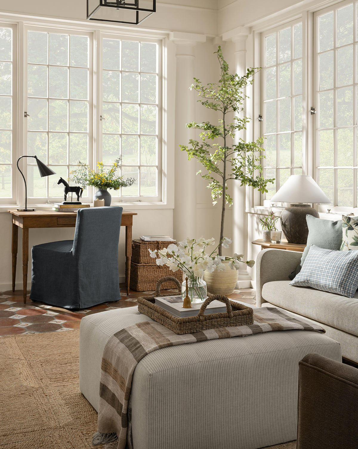 A bright, airy living room with large windows, a beige sofa, a Rowe Fine Furniture Culpepper Ottoman with tray, a small desk and blue chair, plus decorative plants and flowers throughout. McGee & Co.